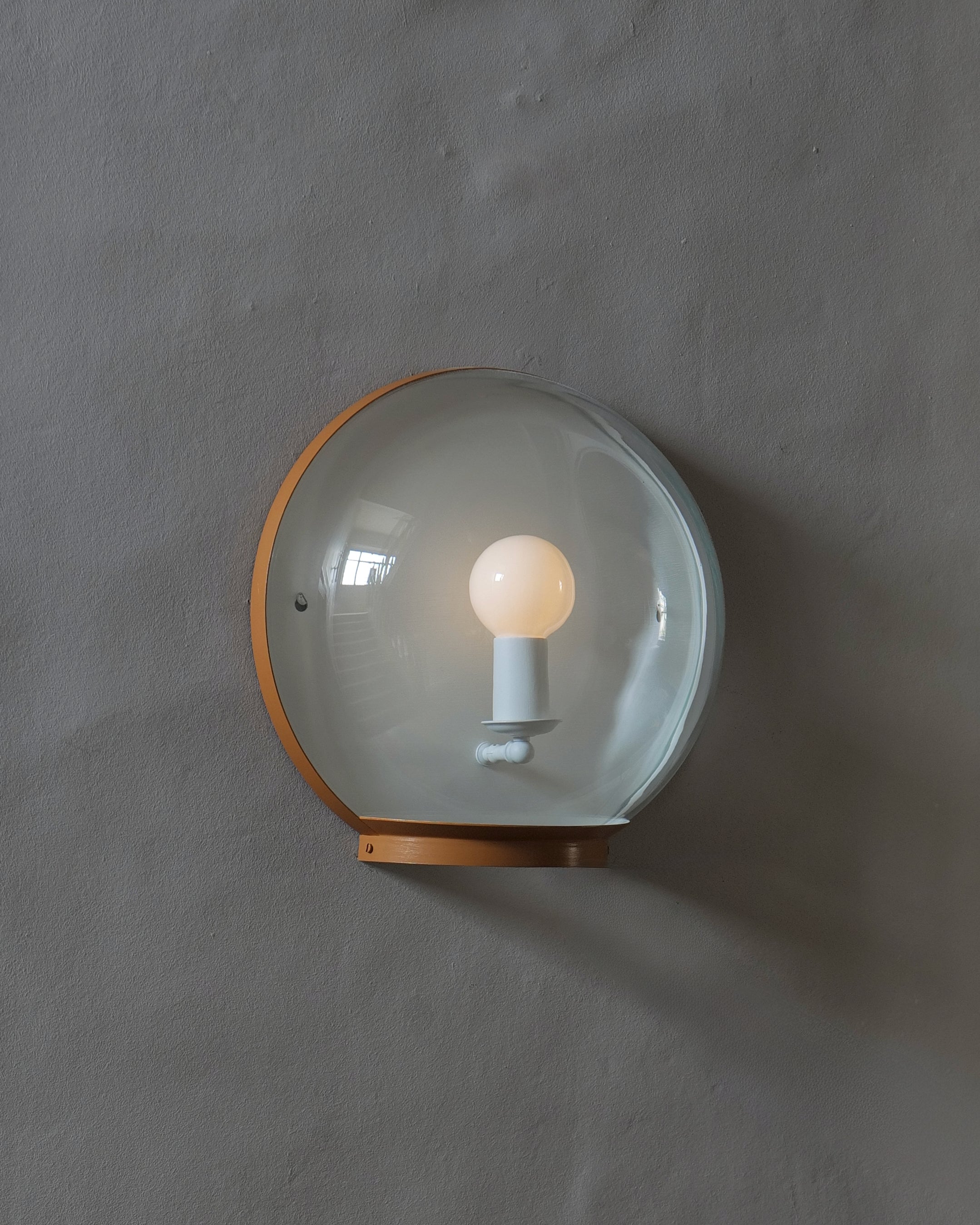 A wall-mounted light fixture with a clear glass dome shade and a warm painted, metallic frame, shown lit against a textured grey wall.