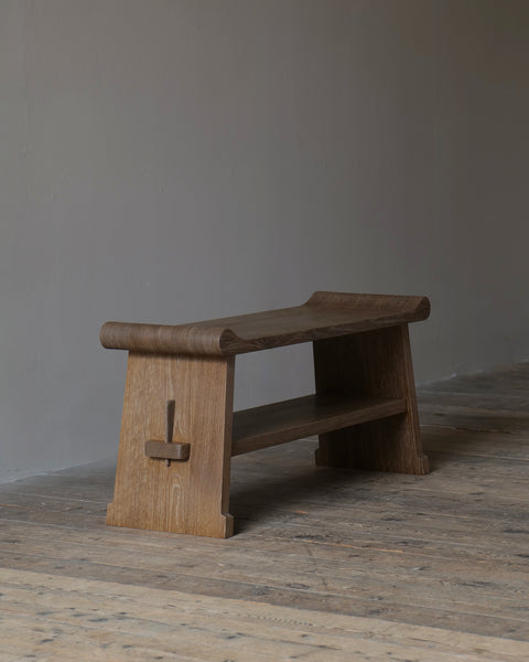 A rustic wooden bench with a curved top and open lower shelf, photographed in a minimalist room with a gray wall and weathered wood floor.