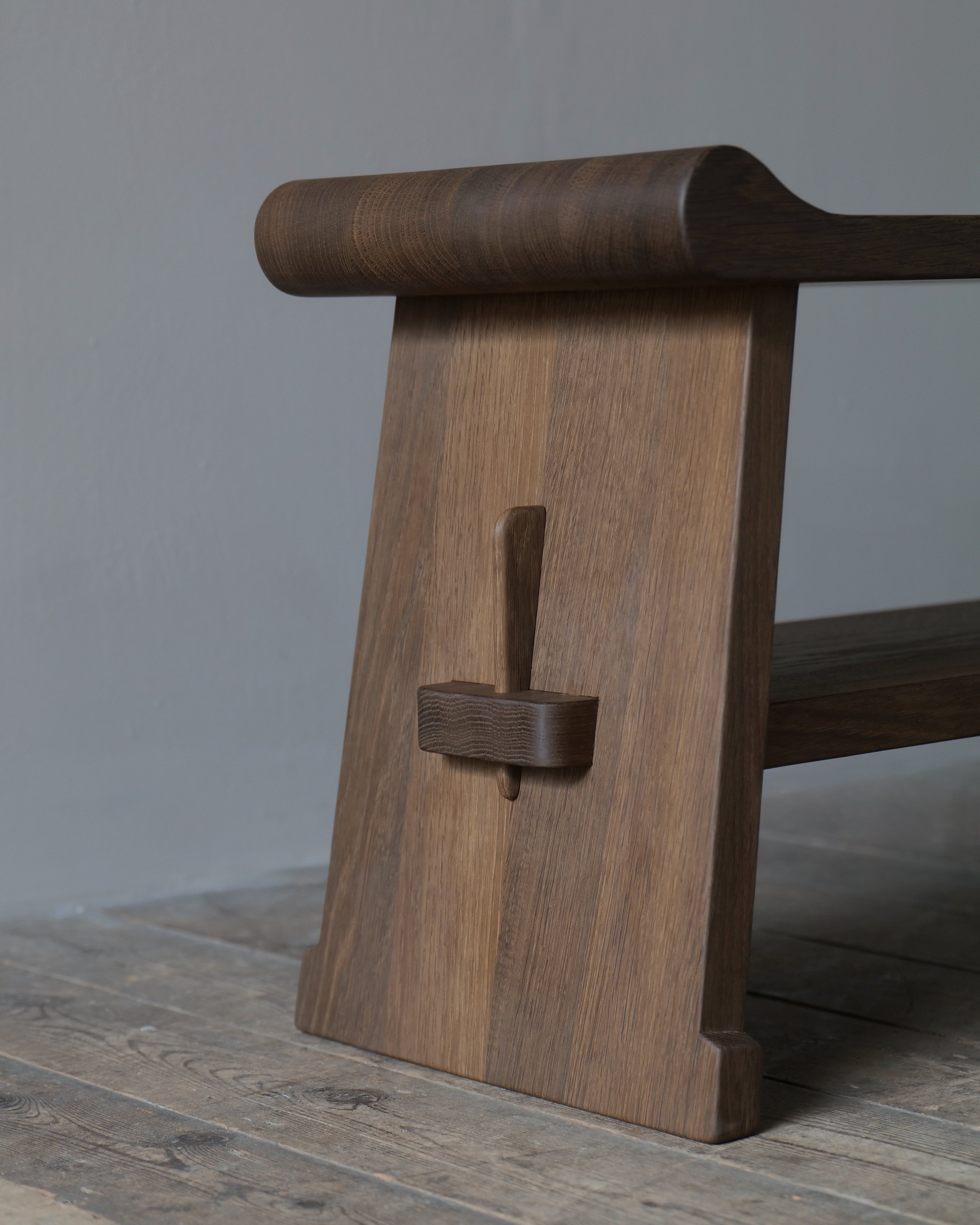 A close-up of an oak bench leg with a curved detail, set on a rustic wooden floor against a neutral grey background.