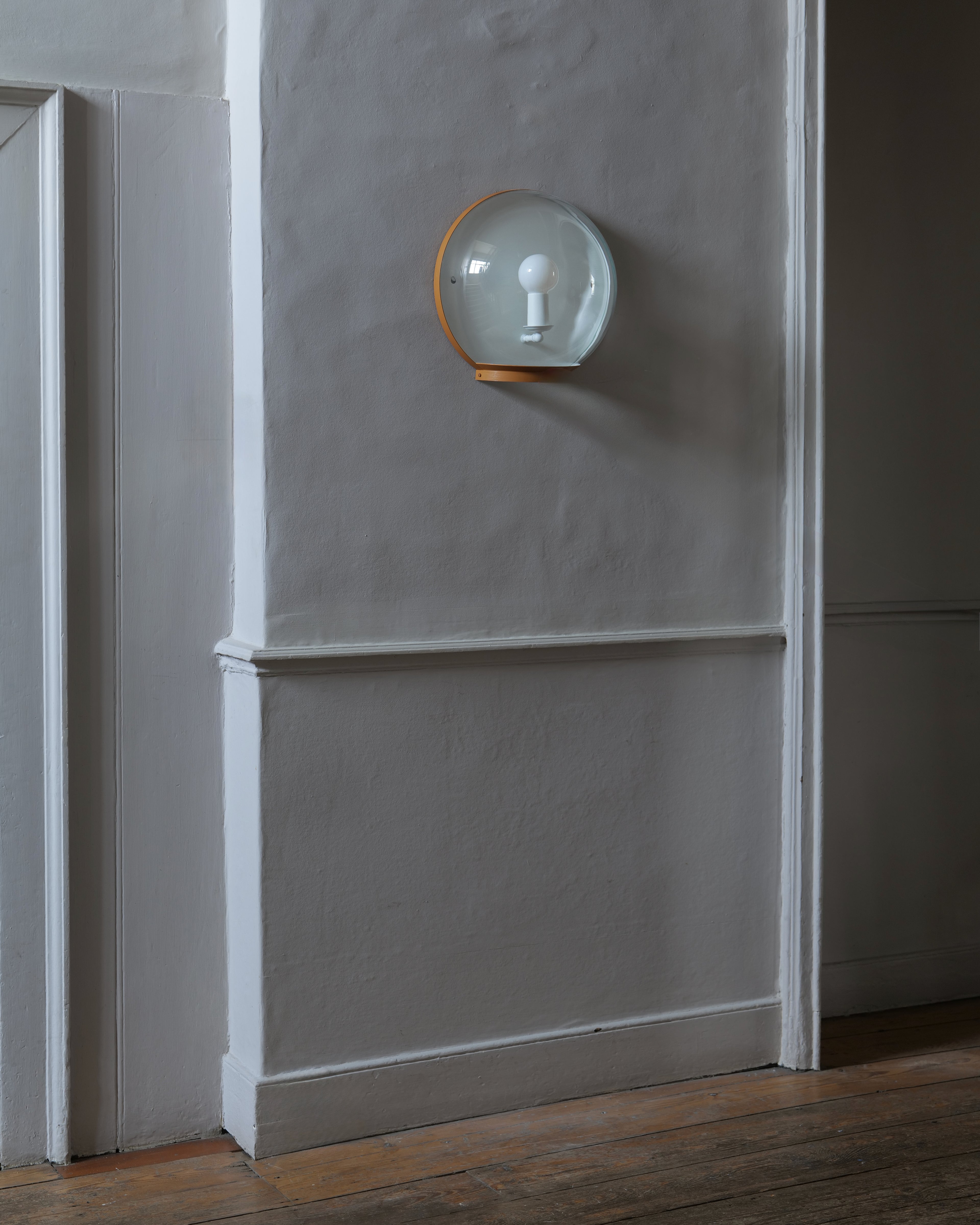 A wall-mounted light fixture with a clear glass dome and a painted metal rim, mounted on a textured grey wall with white trim and a wooden floor.