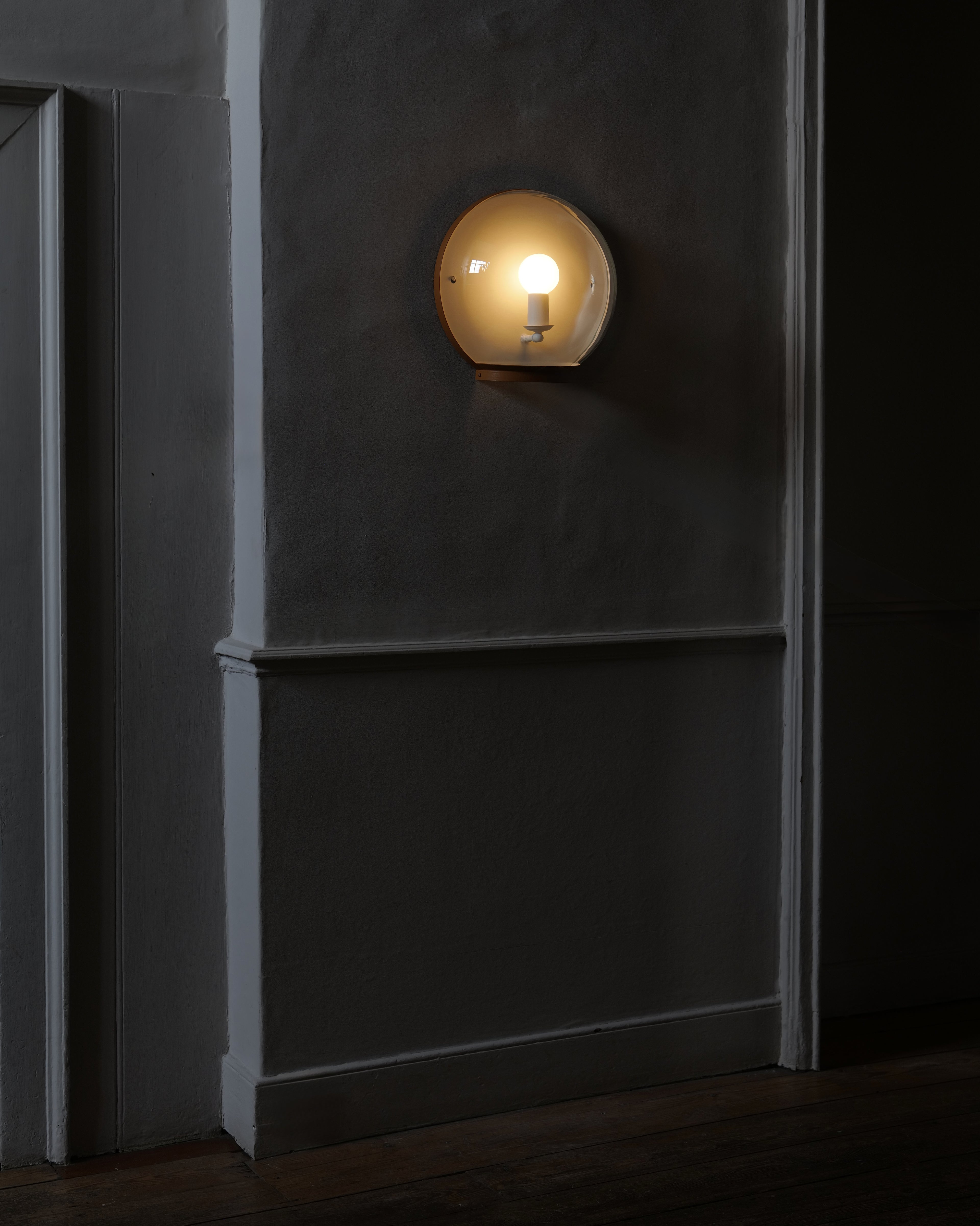 A wall-mounted light fixture with a clear glass dome and a painted metal rim, mounted on a textured grey wall with white trim and a wooden floor. Displayed lit with a warm light.