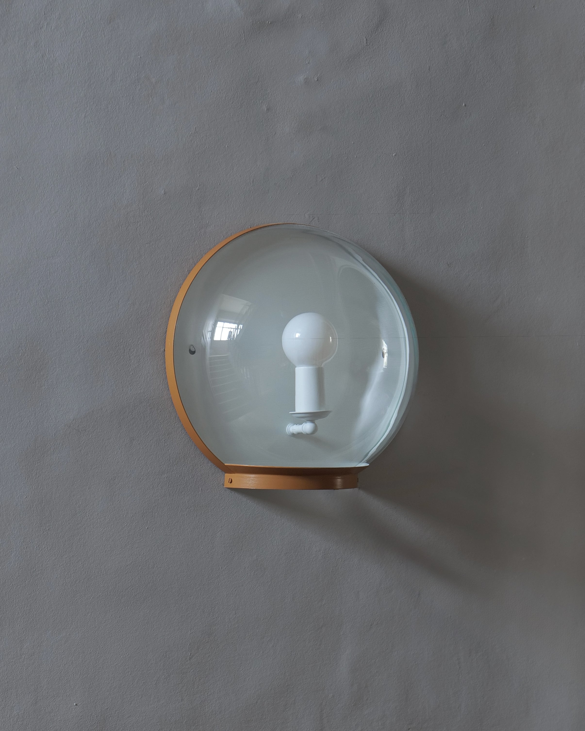 A wall-mounted light fixture with a clear glass dome shade and a warm painted, metallic frame, mounted on a textured grey wall.