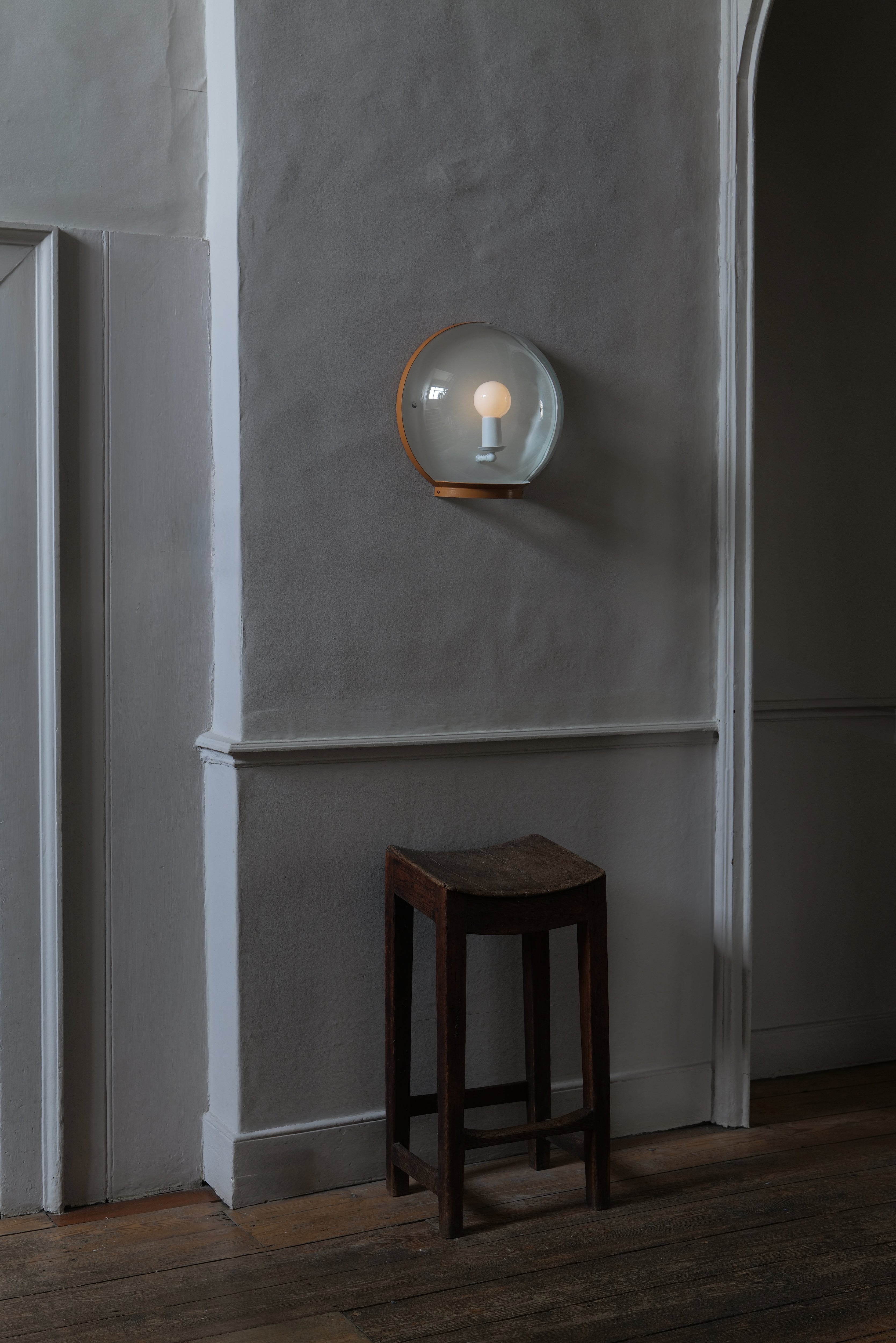A wall-mounted light fixture with a clear glass dome and a painted metal rim, mounted on a textured grey wall, above a dark wooden stool in an interior room.