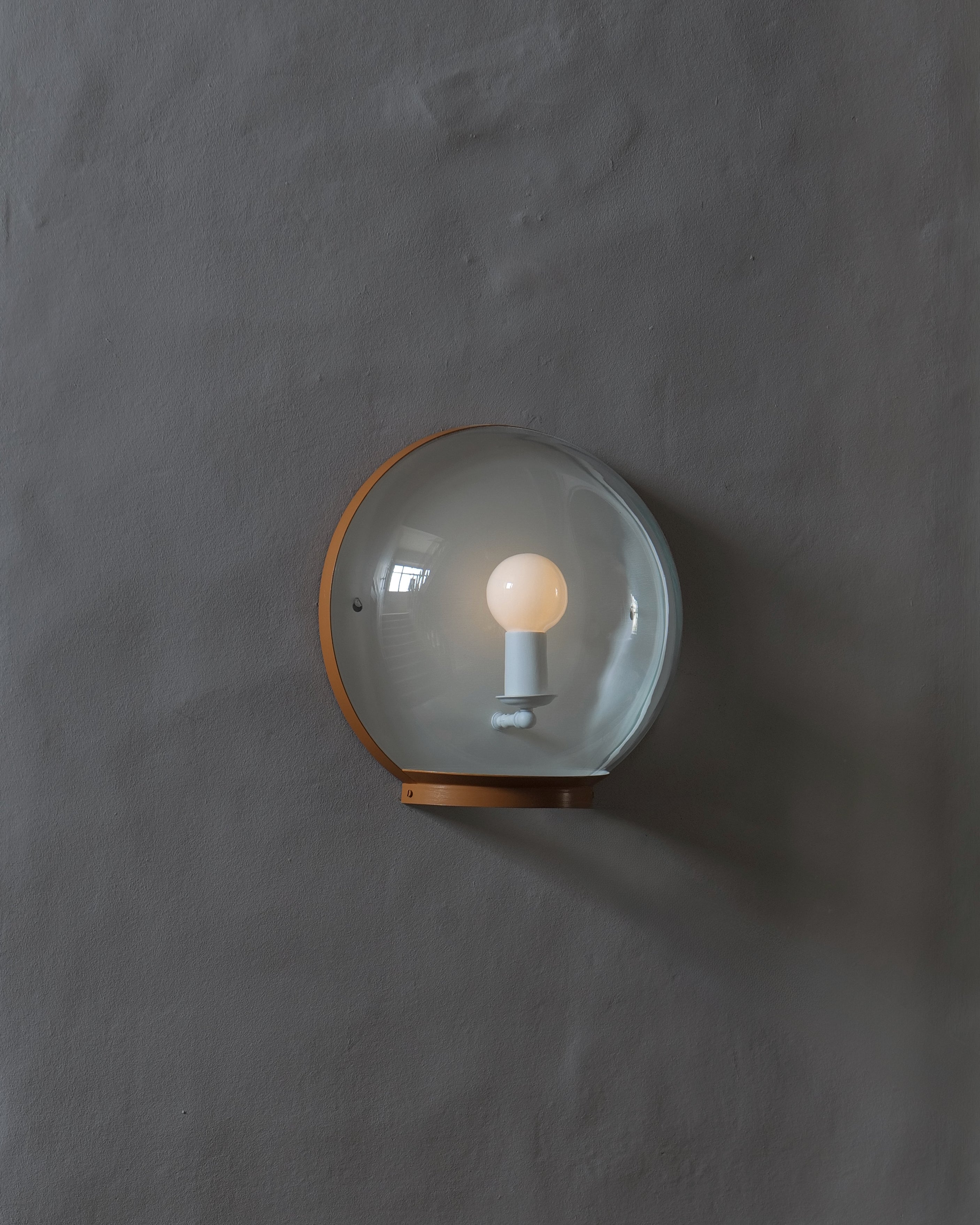 A wall-mounted light fixture with a clear glass dome shade and a warm painted, metallic frame, shown lit against a textured grey wall.