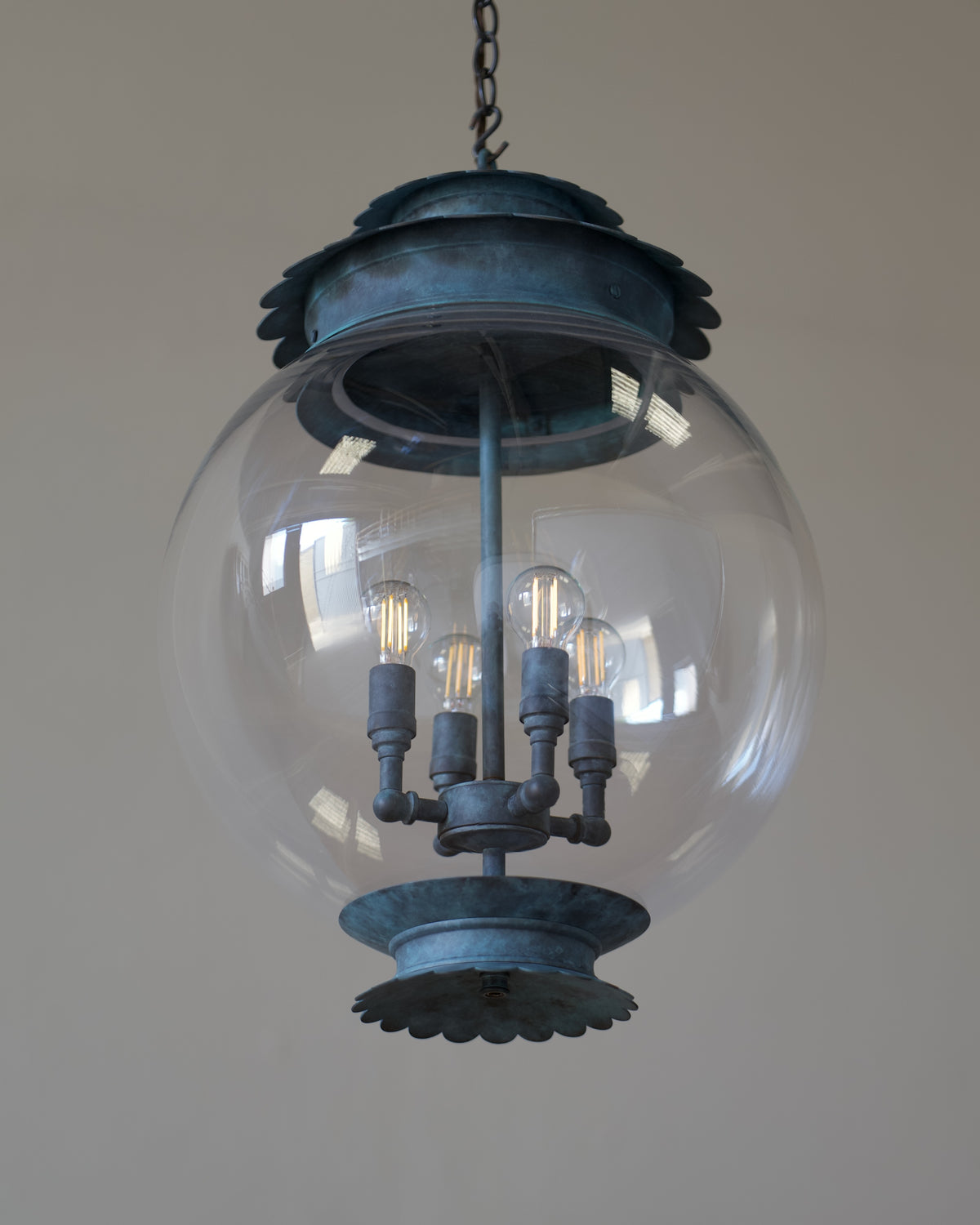 A hanging globe pendant light with a large clear glass sphere and distressed verdigris hardware, shown against a plain beige wall.
