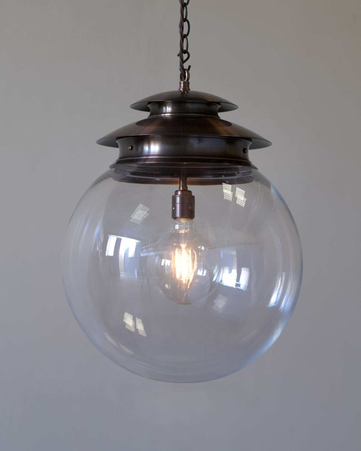 A hanging domus globe lantern with a large clear glass globe shade and a dark bronze metal cap and chain, shown against a plain light grey wall.