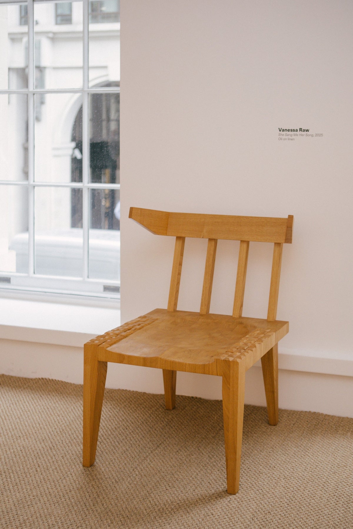 A light natural wood armchair with a slatted backrest and seat, positioned on a beige woven rug against a white wall with a window.