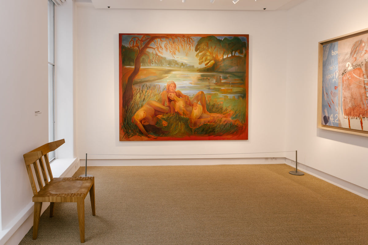 A large framed painting hangs on a white gallery wall in a museum or art gallery. The painting depicts a warm-toned outdoor scene with figures near water and trees.