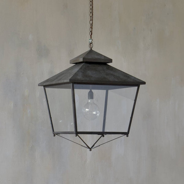 A dry bronze brass lantern-style pendant light hangs from a chain against a textured grey wall and a light wood floor.