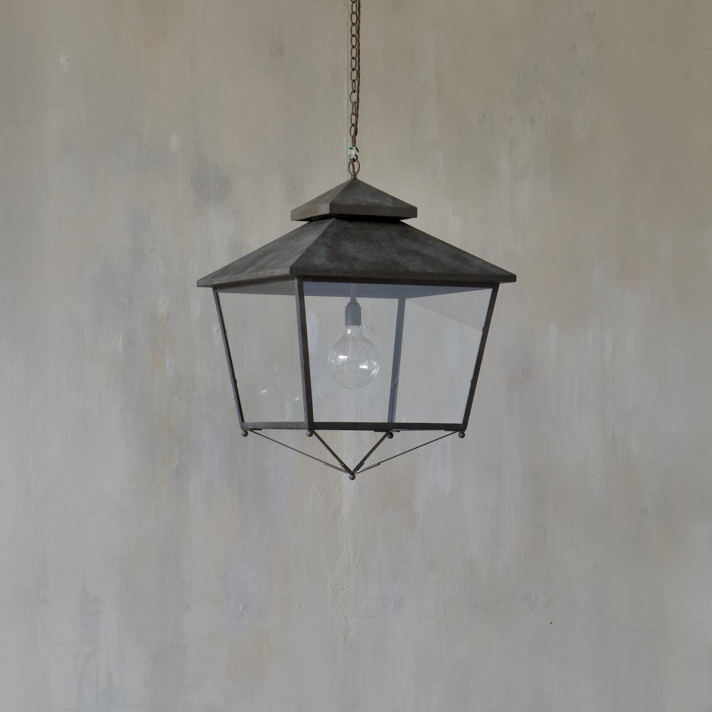 A dry bronze brass lantern-style pendant light hangs from a chain against a textured gray wall and light wood floor.