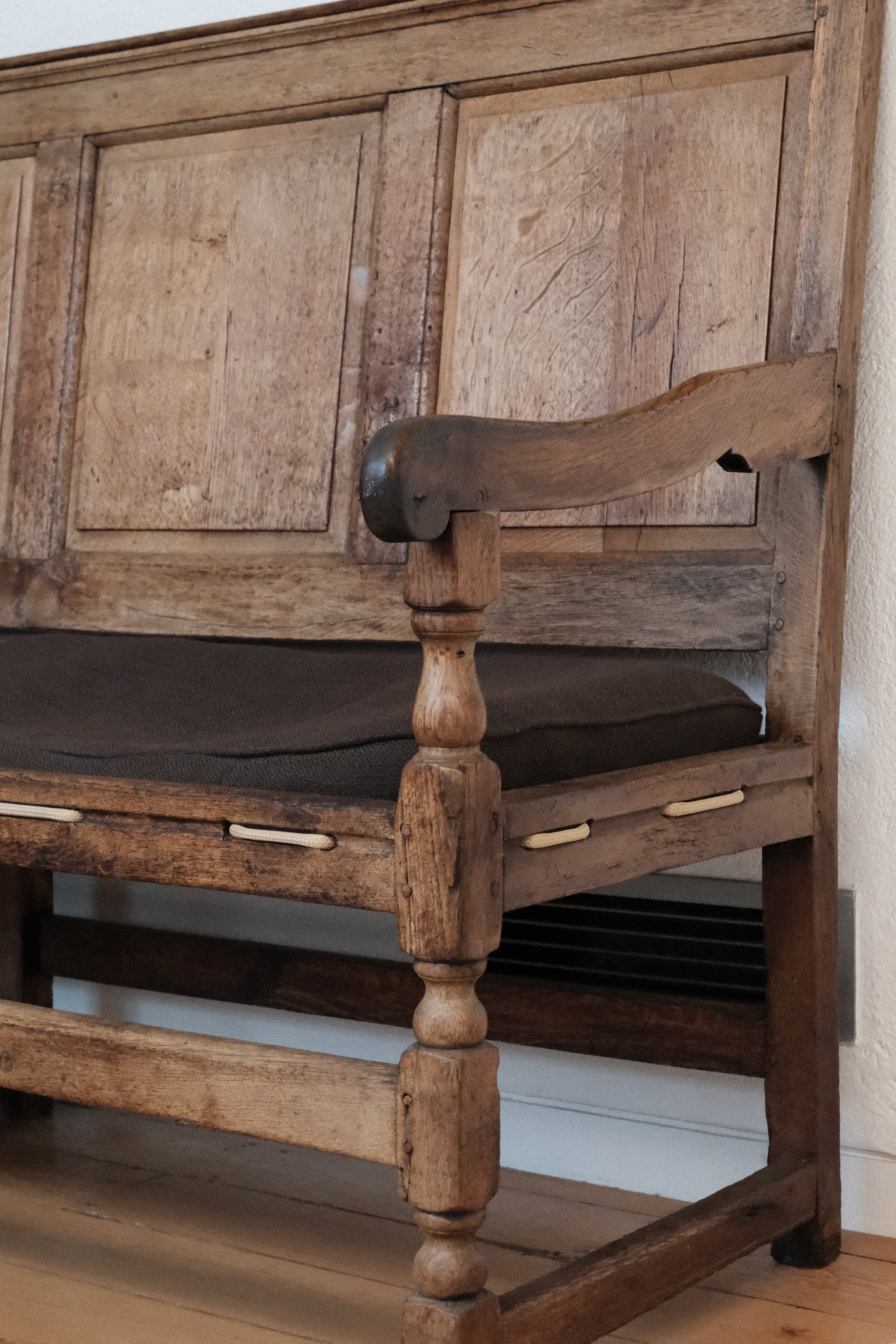 A rustic wooden bench with a paneled backrest, turned spindle legs, and a dark cushion, shown in a close-up indoor shot.