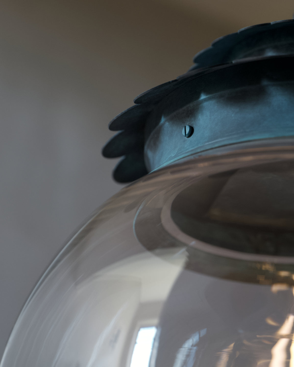Close-up detail of a transparent glass globe with a verdigris roof and a small visible screw fastener, photographed indoors against a grey wall.