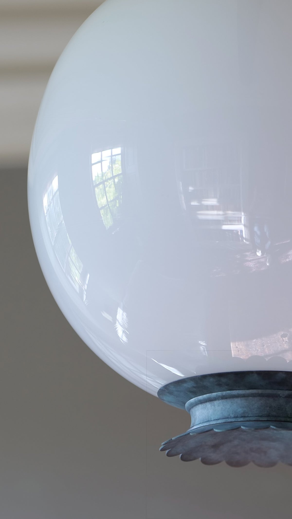 A close-up of a large, glossy white glass globe shade attached to a verdigris finial fixture, set against a grey background.