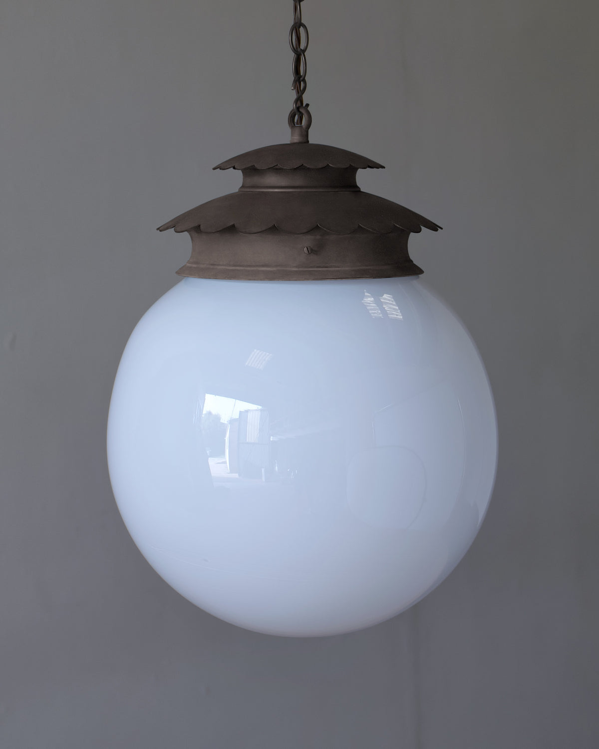 A vintage-style pendant light with a large, opaque white glass globe shade and a sry bronze metal cap and chain, hanging against a plain gray wall.