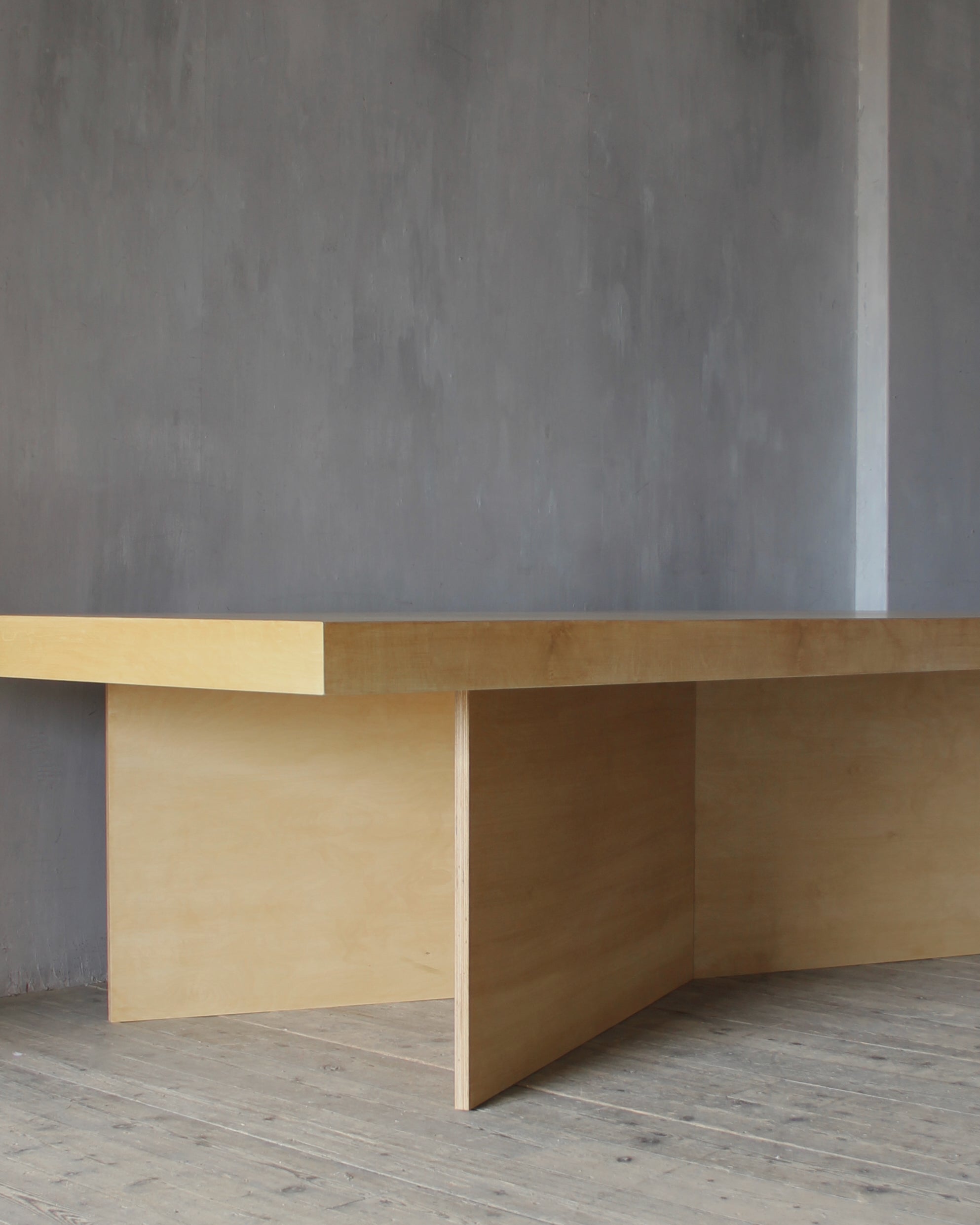 A minimalist light wood table with a thick rectangular top and integrated side panels, shown in a room with a textured gray wall and wood floor.
