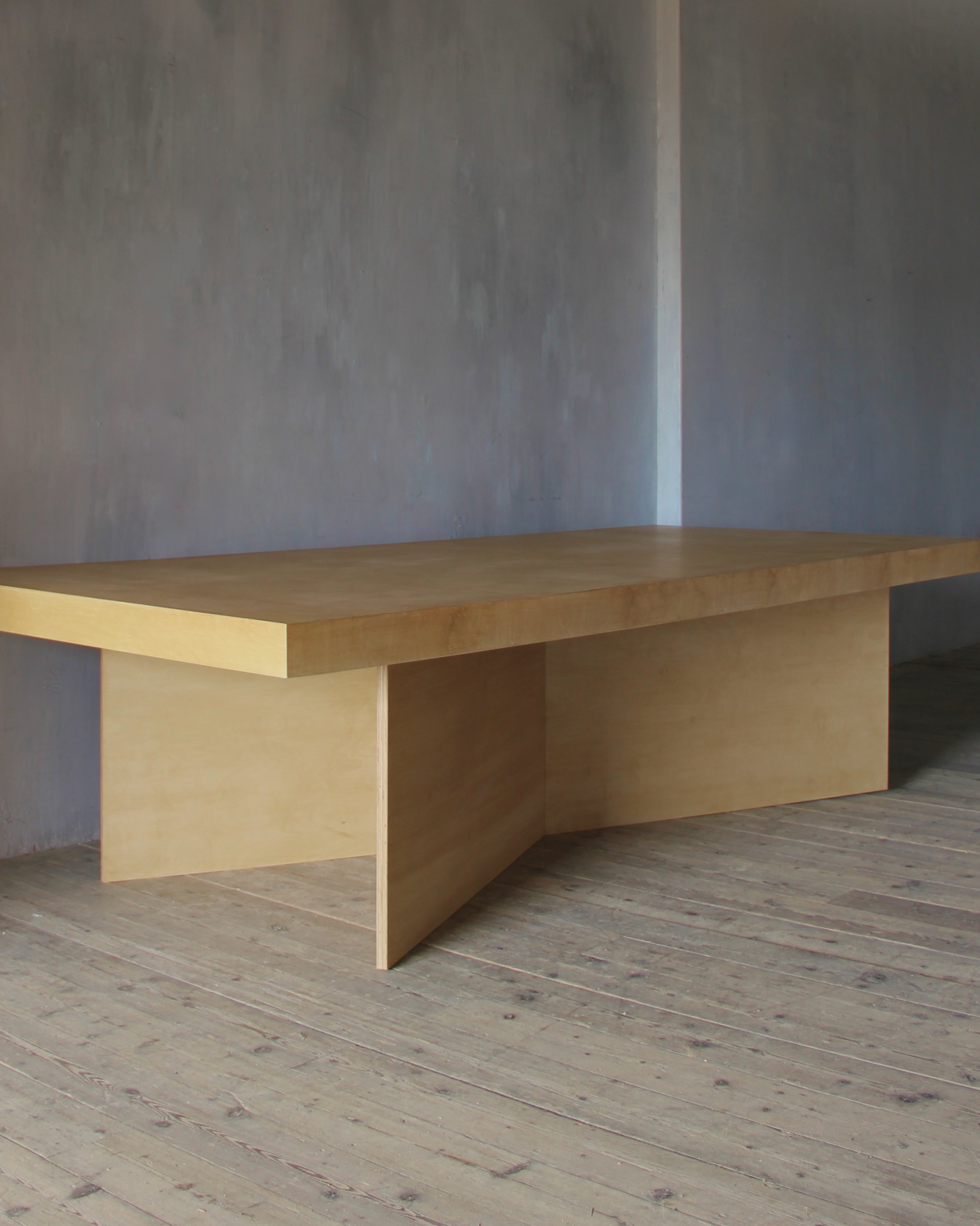 A minimalist light wood conference table with a thick rectangular top and integrated pedestal base, set against a textured gray wall and wooden floor.
