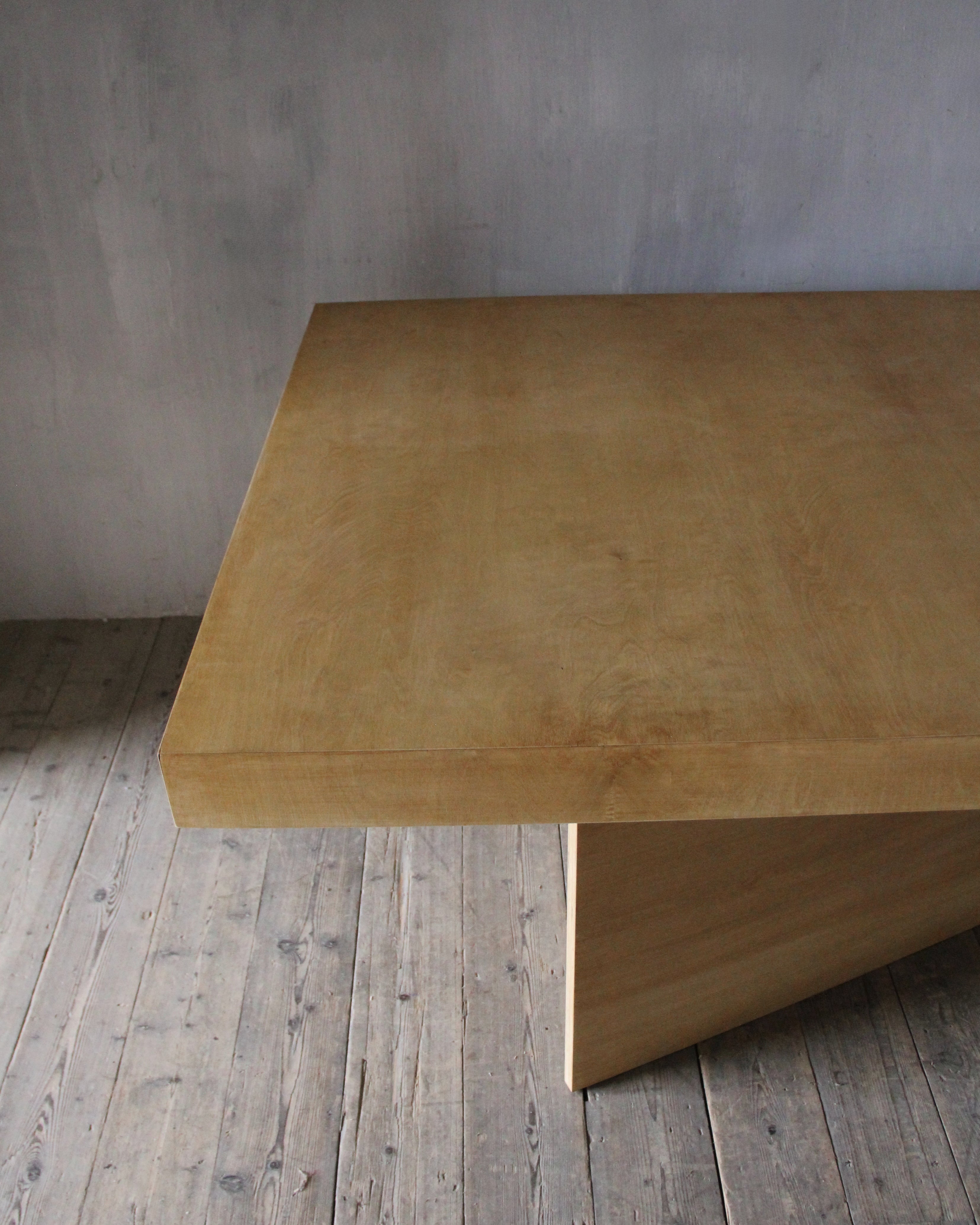 A minimalist light wood table with a thick rectangular top and a single visible block-style leg, photographed in a rustic room with a gray wall and weathered wood floor.