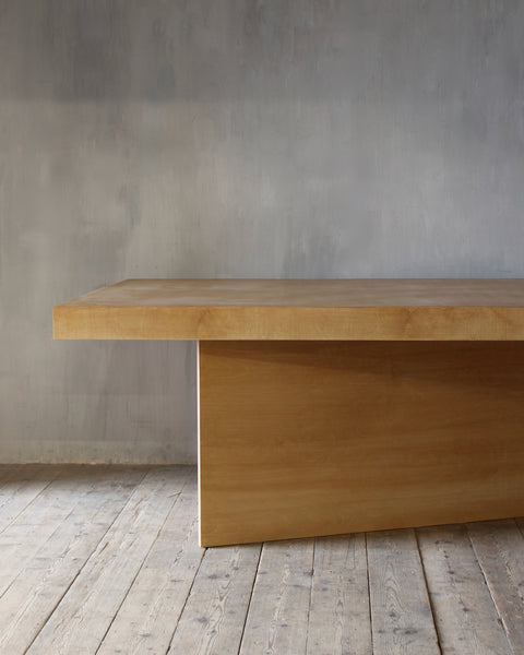 A minimalist light wood table with a thick rectangular top and a single block base, set against a textured gray wall and light wood plank floor.