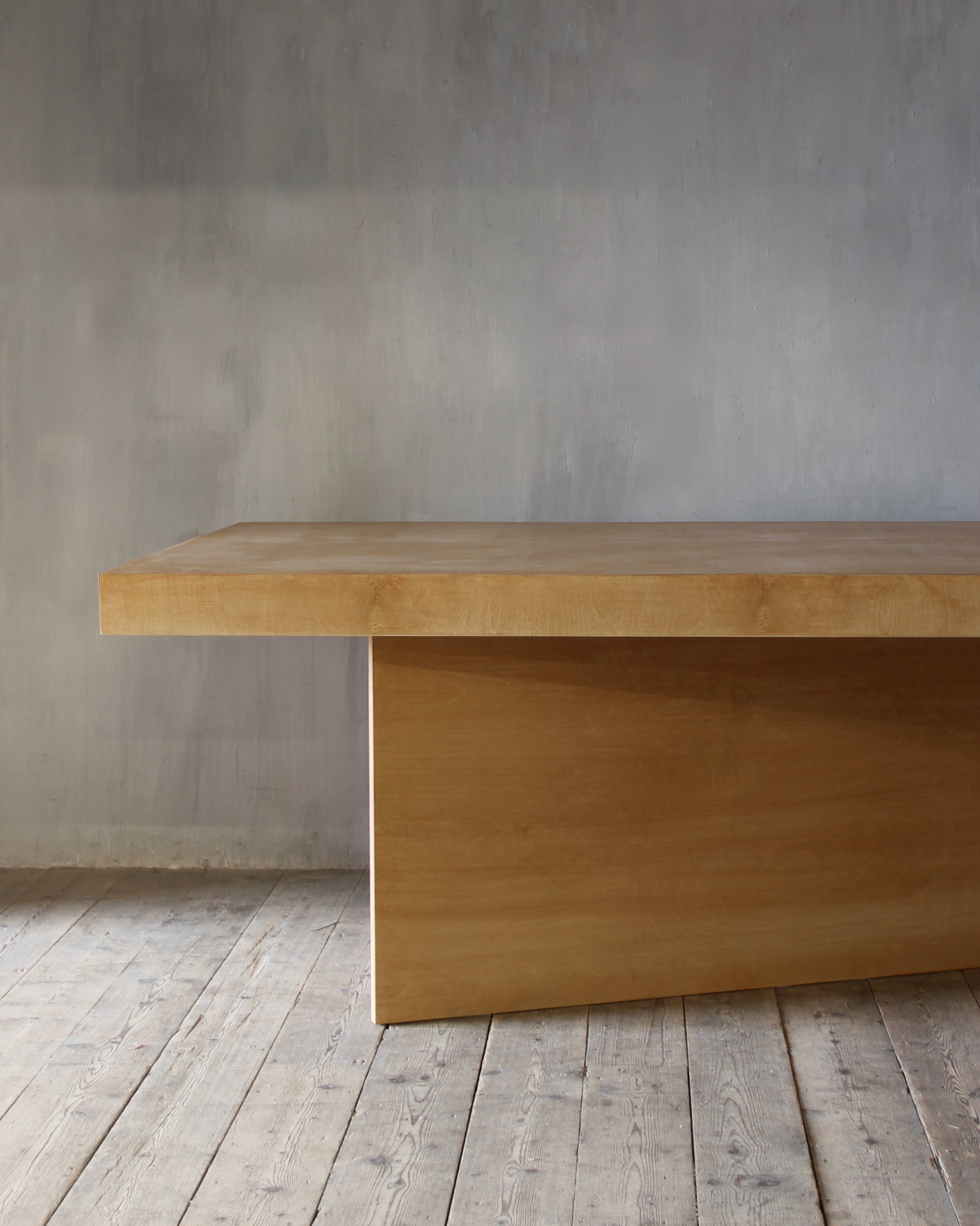 A minimalist light wood table with a thick rectangular top and a single block base, set against a textured gray wall and light wood plank floor.