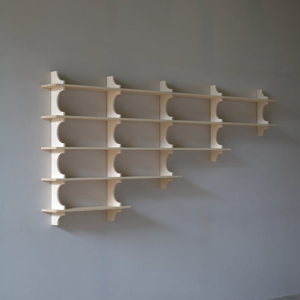 A set of light natural wood wall shelves mounted on a grey wall above a wooden floor.