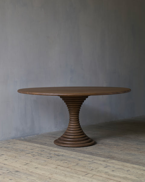 A circular, oak dining table with a sculptural, ribbed pedestal base, set against a textured grey wall and light wood floor.