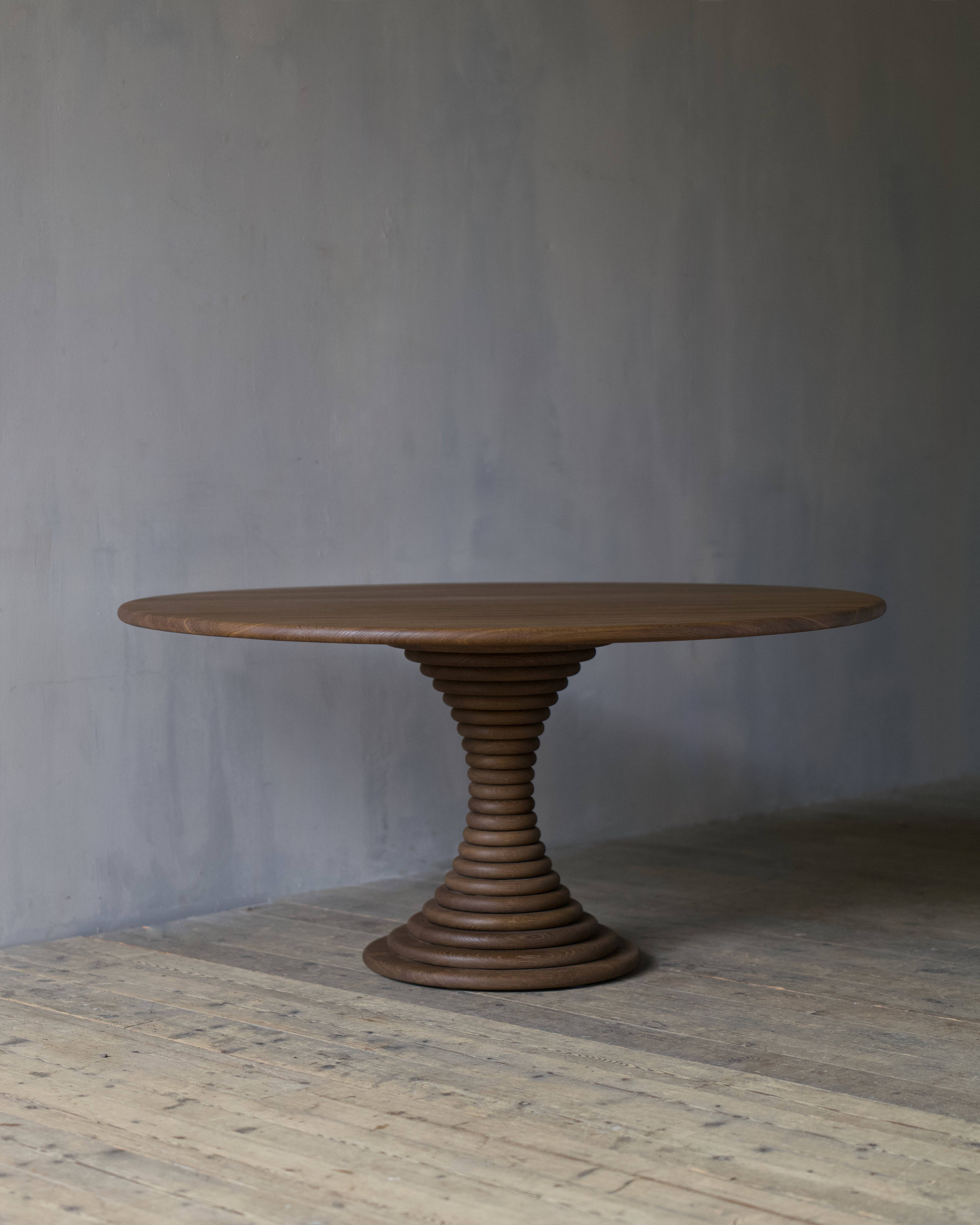 A circular, oak dining table with a sculptural, ribbed pedestal base, set against a textured grey wall and light wood floor.