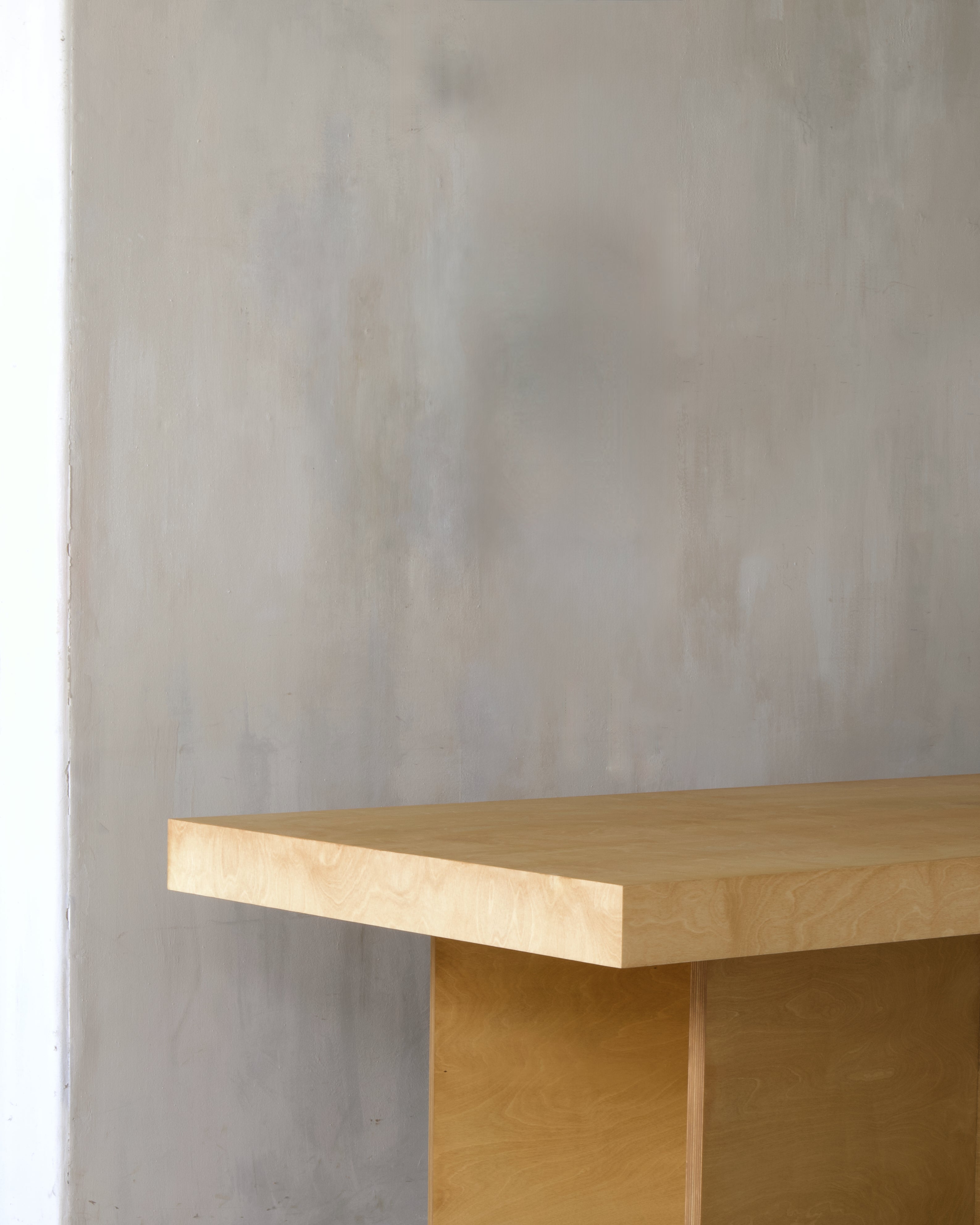 A close-up of a light natural wood table or desk corner against a textured gray plaster wall.
