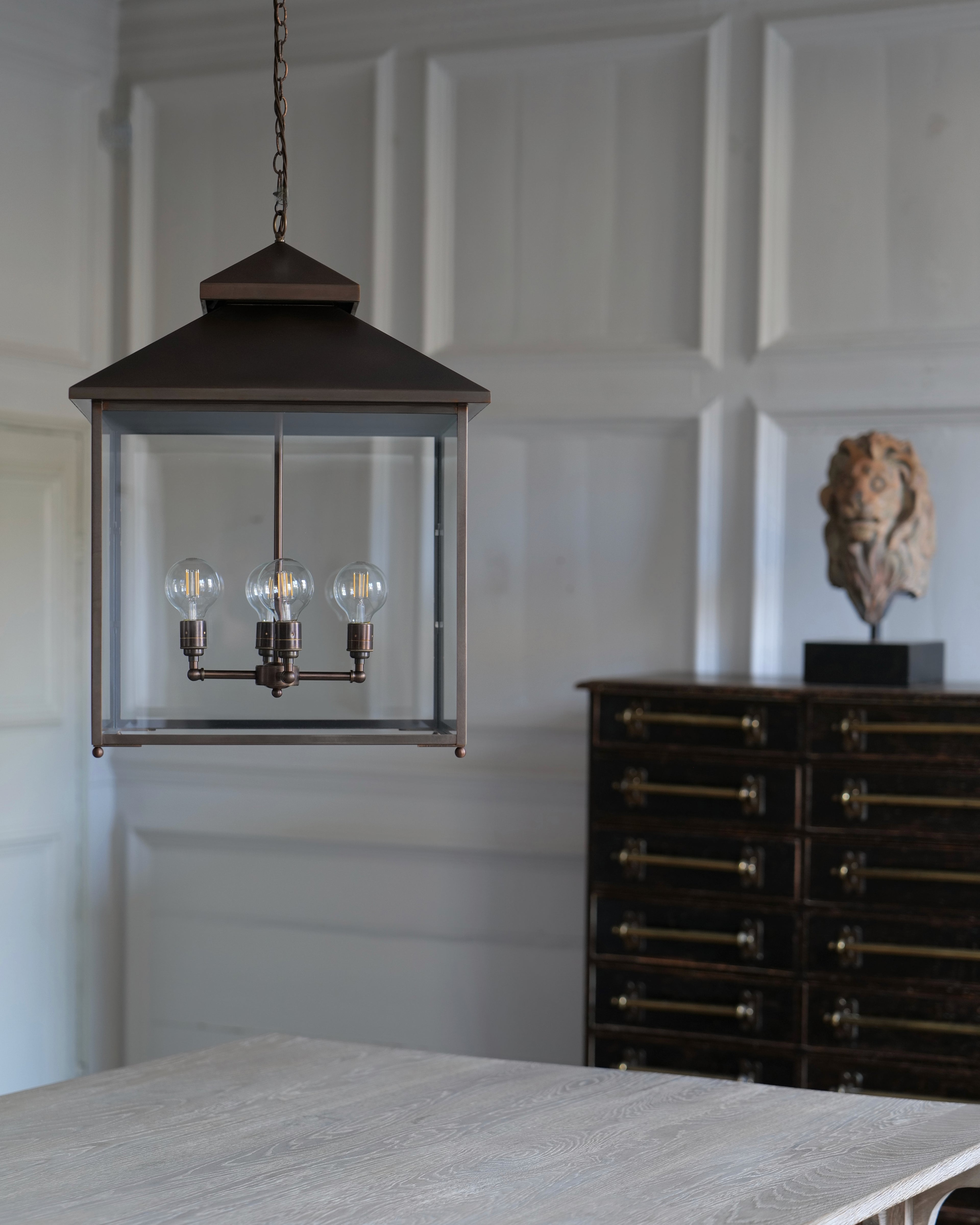 A dark brass lantern-style pendant light with clear glass panels and three visible bulb filaments, hanging in a room with white panelled walls, a wooden table in the foreground, and a dark wood cabinet in the background.