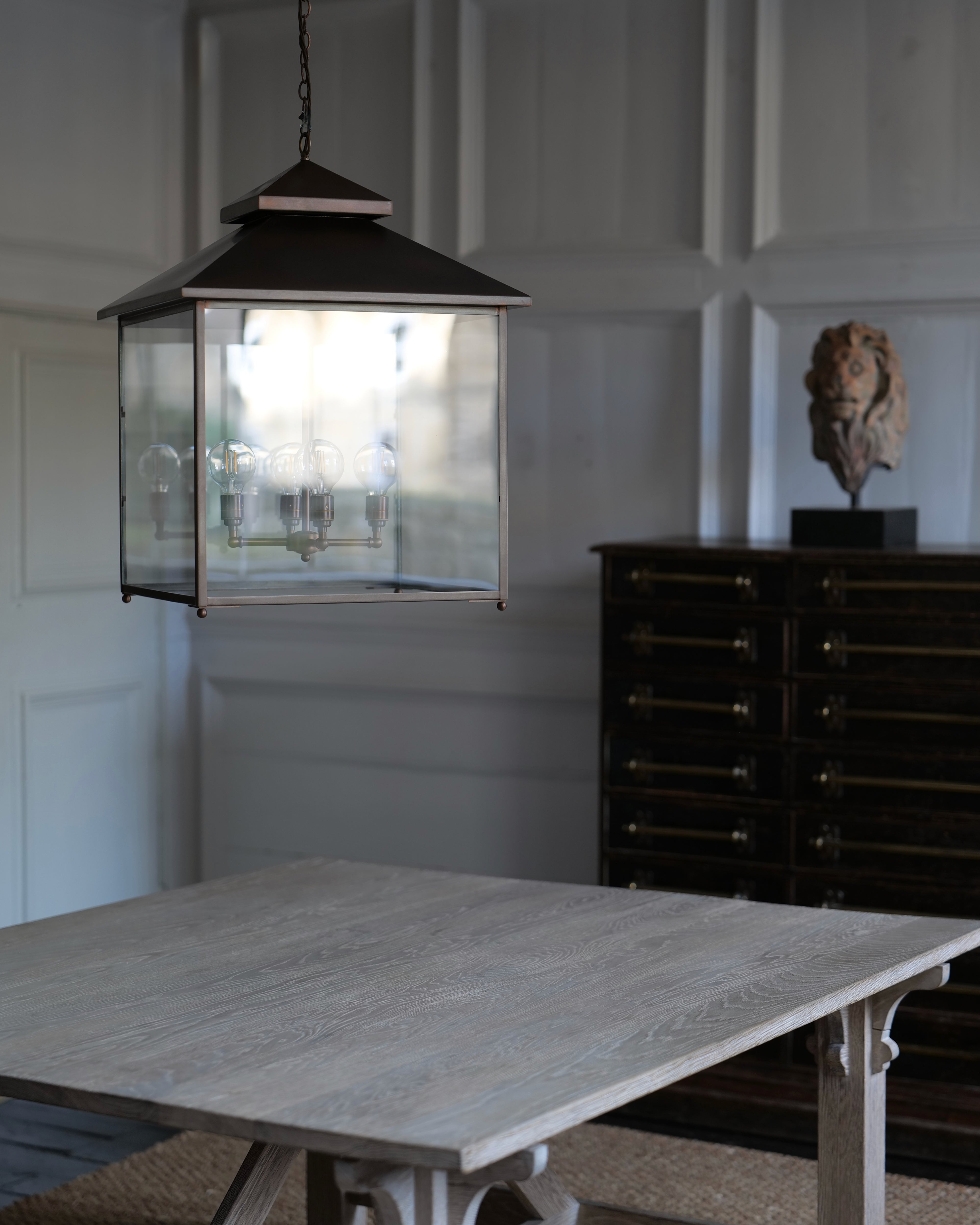 A hanging lantern-style pendant light with a dark metal frame and clear glass panels, suspended above a light wood dining table in a softly lit room with paneled walls and a dark cabinet in the background.