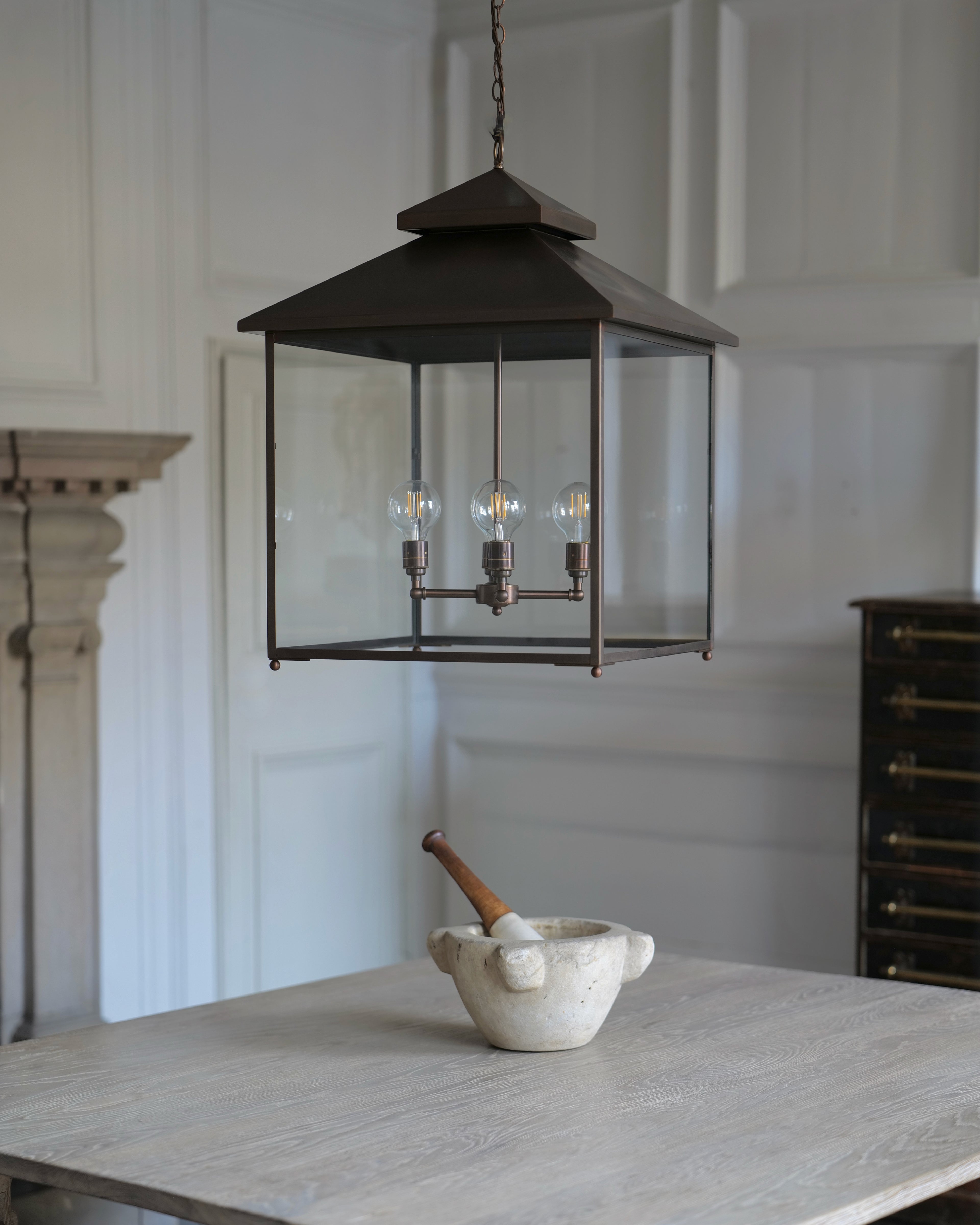 A dark brass lantern-style pendant light with clear glass panels hangs over a light wood table in a bright, white-panelled room. A small white ceramic mortar and pestle sits on the table below.