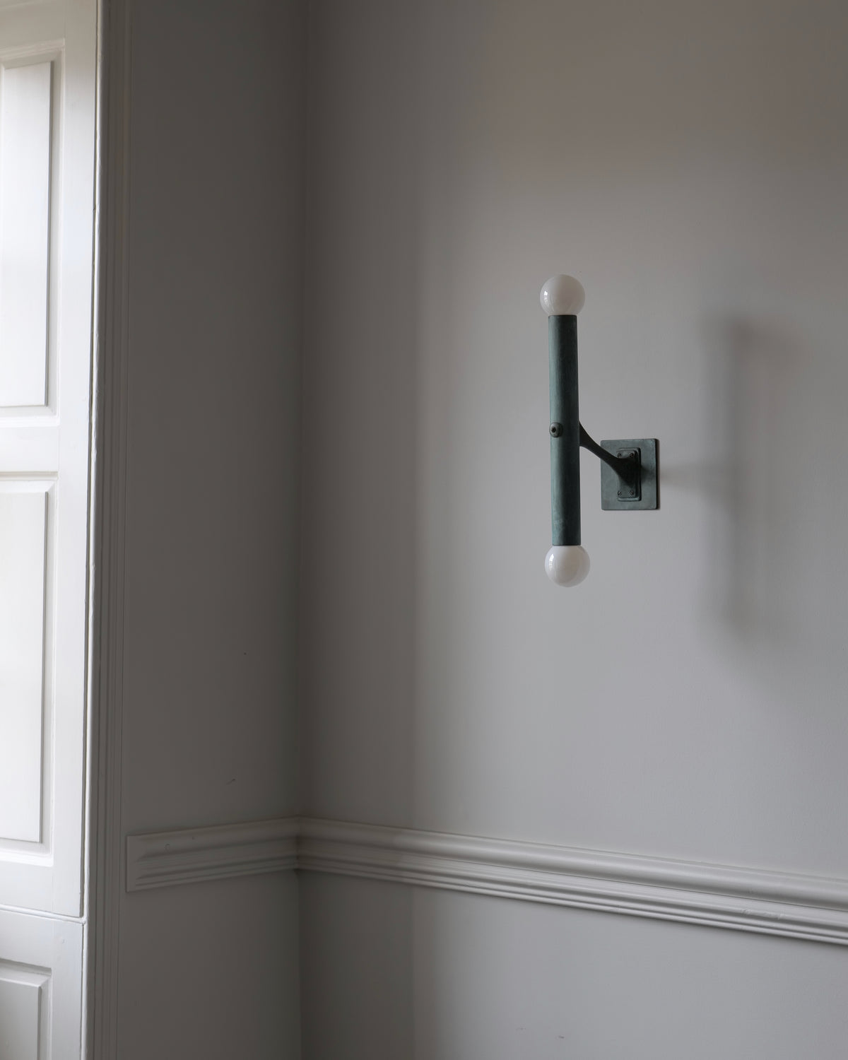 A vesper wall sconce with a verdigris brass frame and two opal spherical glass bulbs, mounted on a light gray wall in an interior setting.