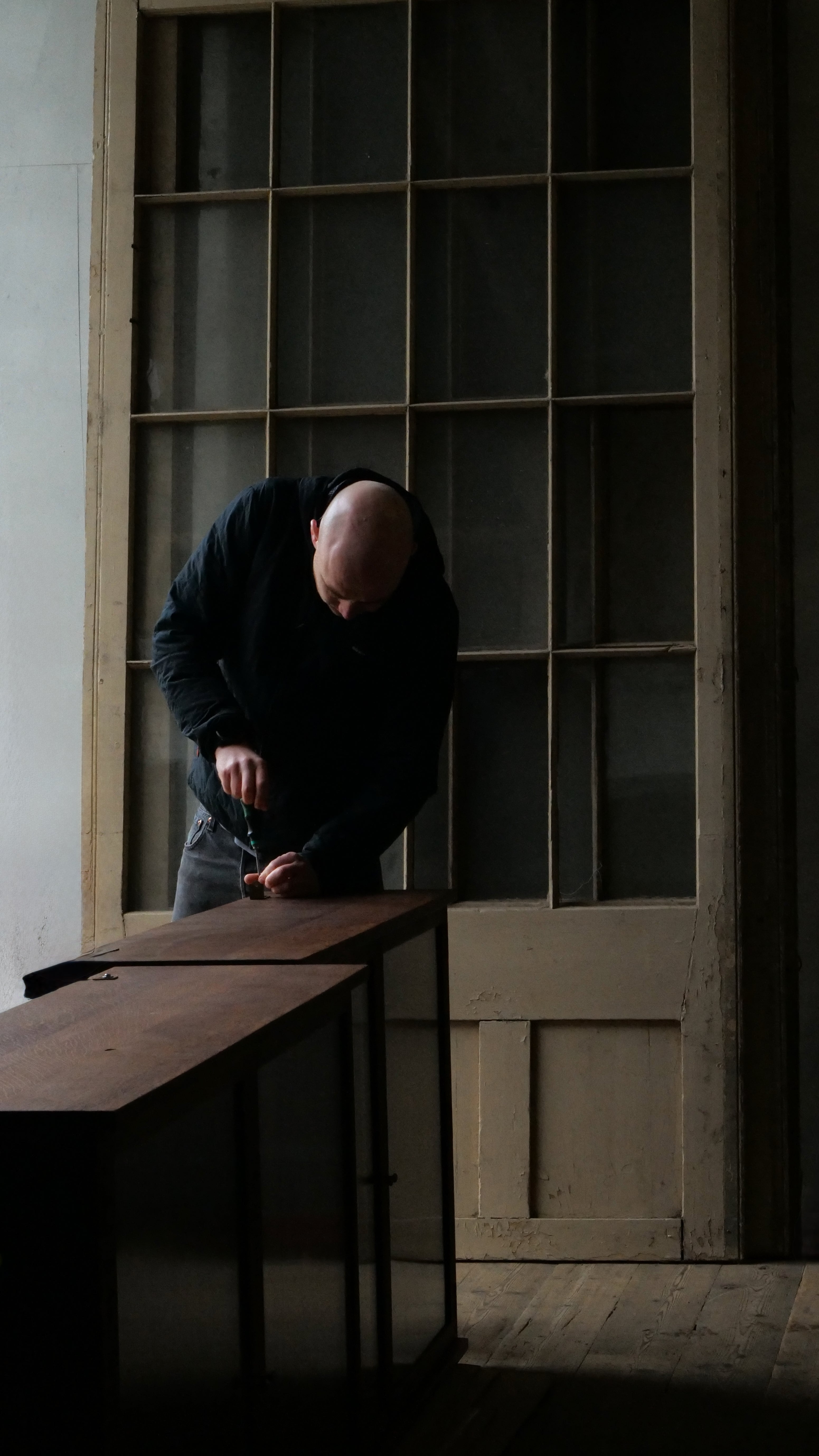 Man restoring a wooden cabinet in a dimly lit room with a large window.