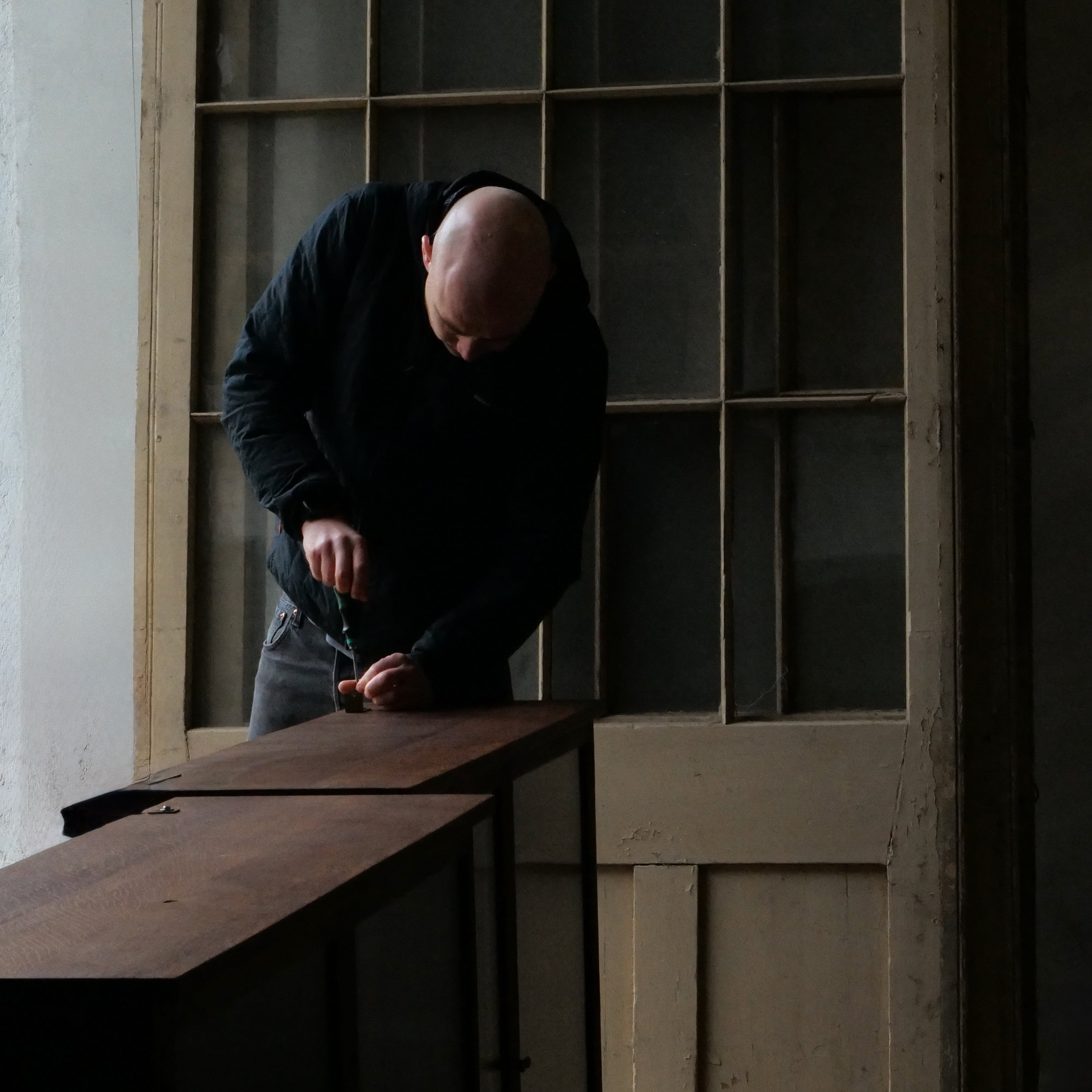 A man restoring a cabinet in a dimly lit room with large windows.
