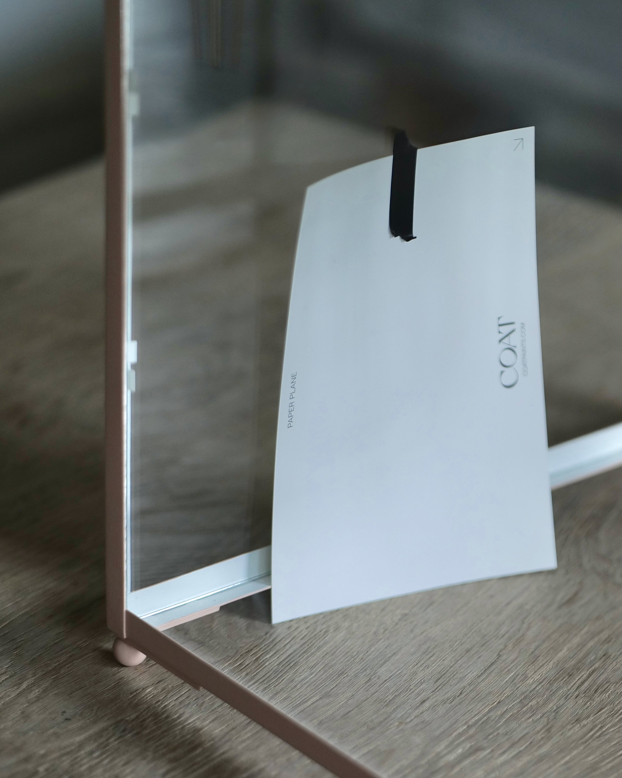 A close-up of a pale pink paint swatch with the brand name 'COAT' printed on it, held by black tape against a clear glass panel on the inside of a lantern, set on a light wood surface.