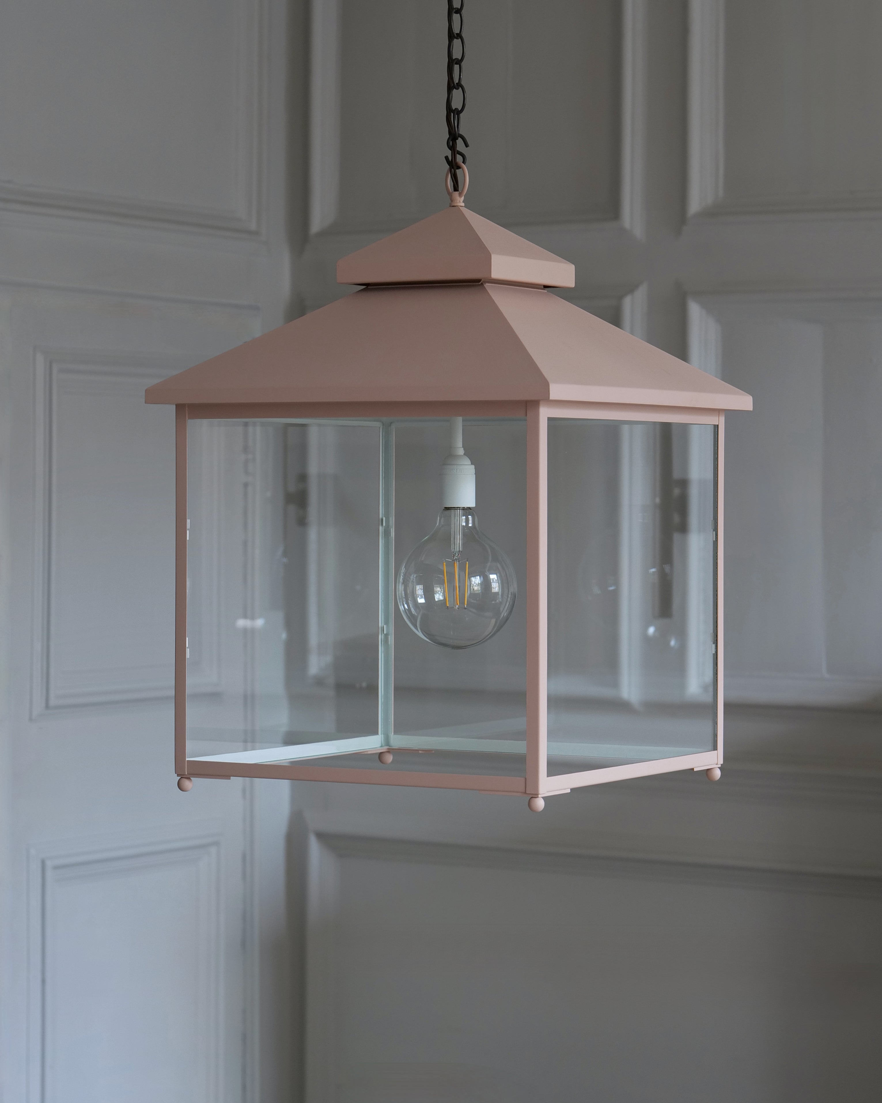 A dusty pink metal lantern-style pendant light with clear glass panels and a visible bulb, hanging from a dark chain against a white-panelled wall.