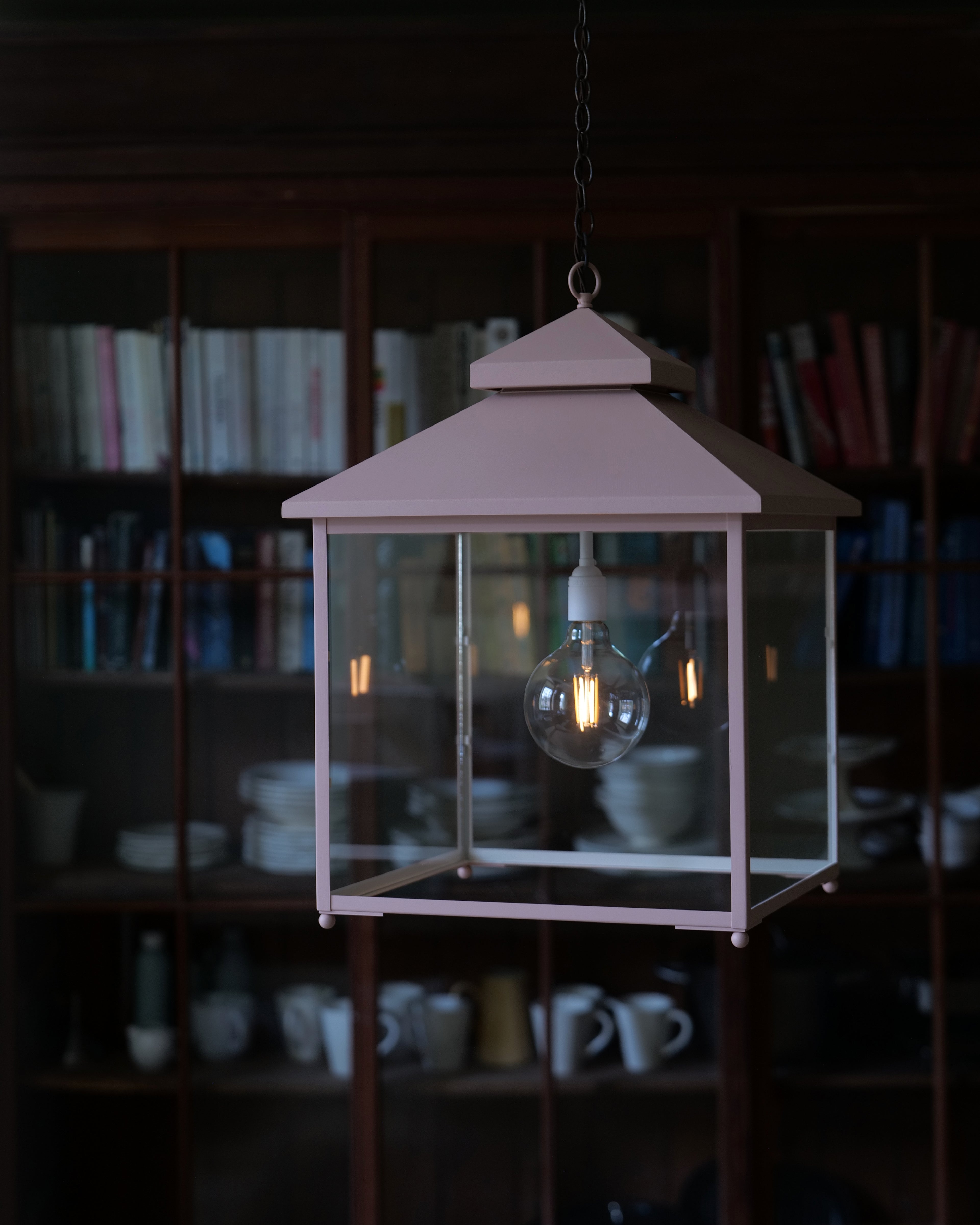 A pink metal lantern-style pendant light with a clear glass front panel and a visible glowing bulb, hanging in front of a dark bookshelf interior.