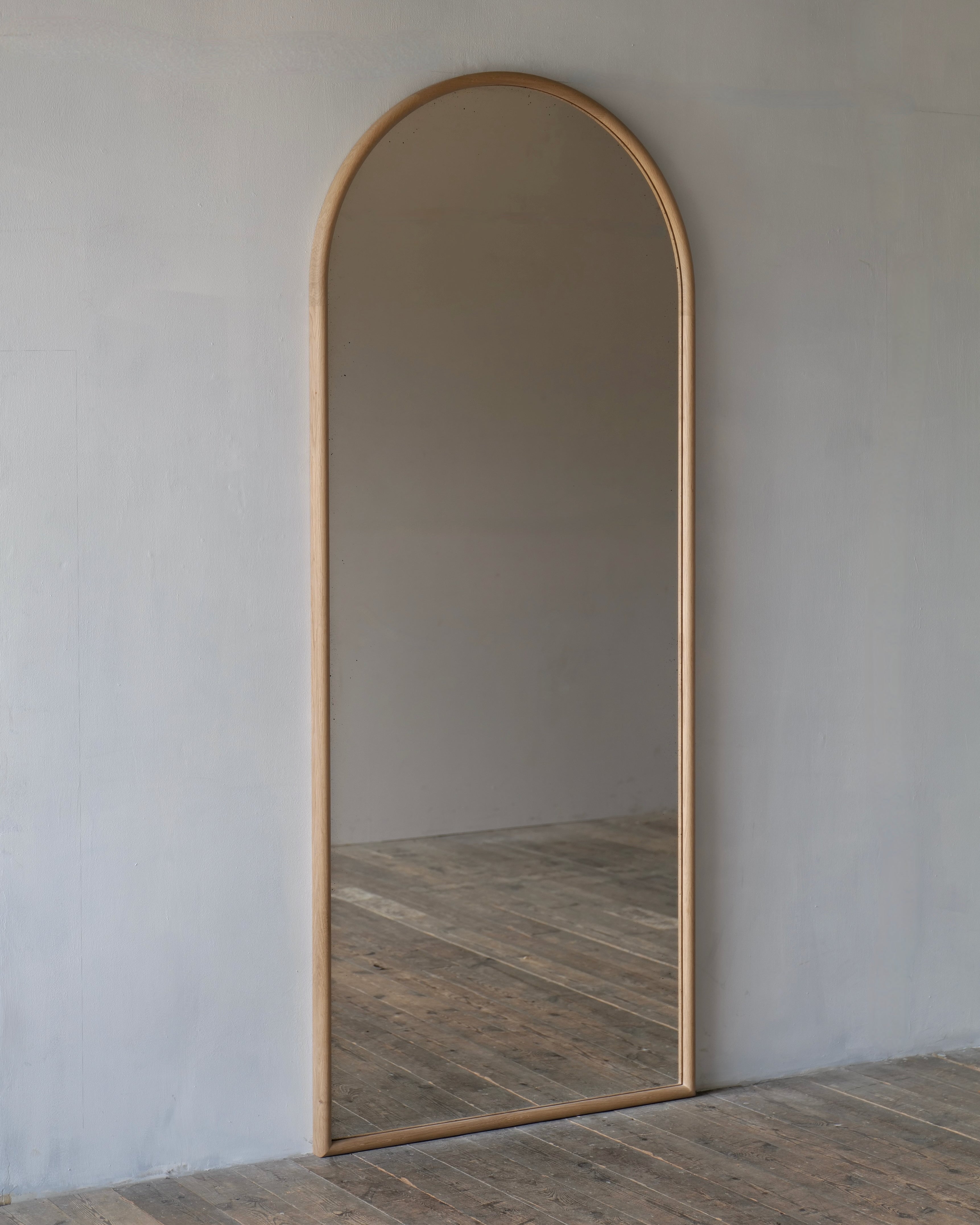An arched full-length floor mirror with a light natural wood frame, leaning against a textured light gray wall on a rustic wooden plank floor.