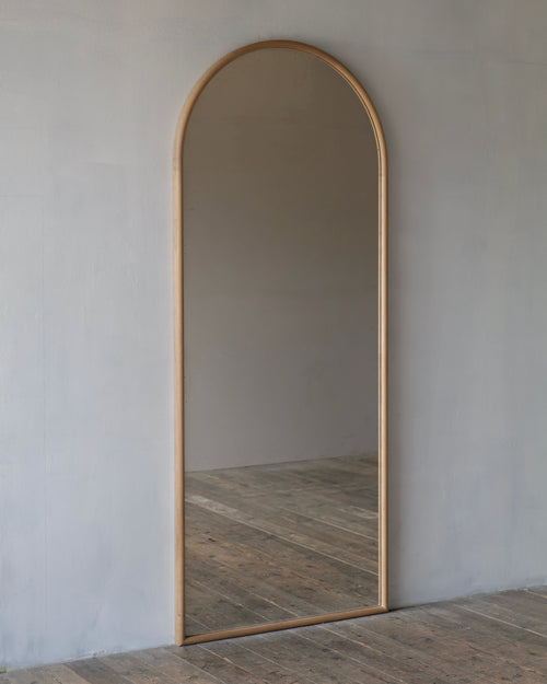 An arched full-length floor mirror with a light natural wood frame, leaning against a textured light gray wall on a rustic wooden plank floor.