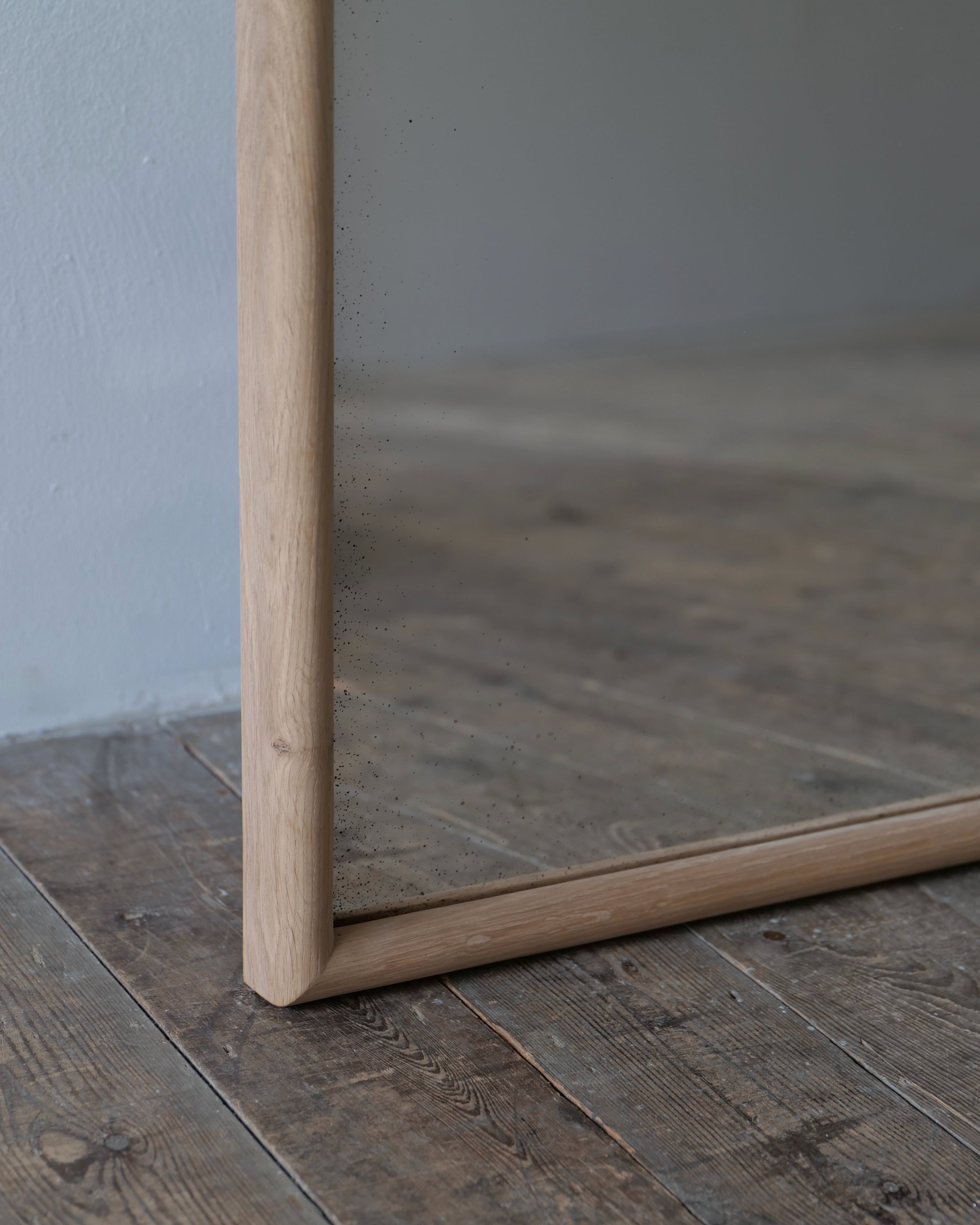 A large rectangular mirror with a natural light wood frame, shown resting on a rustic dark wood plank floor against a neutral gray wall.