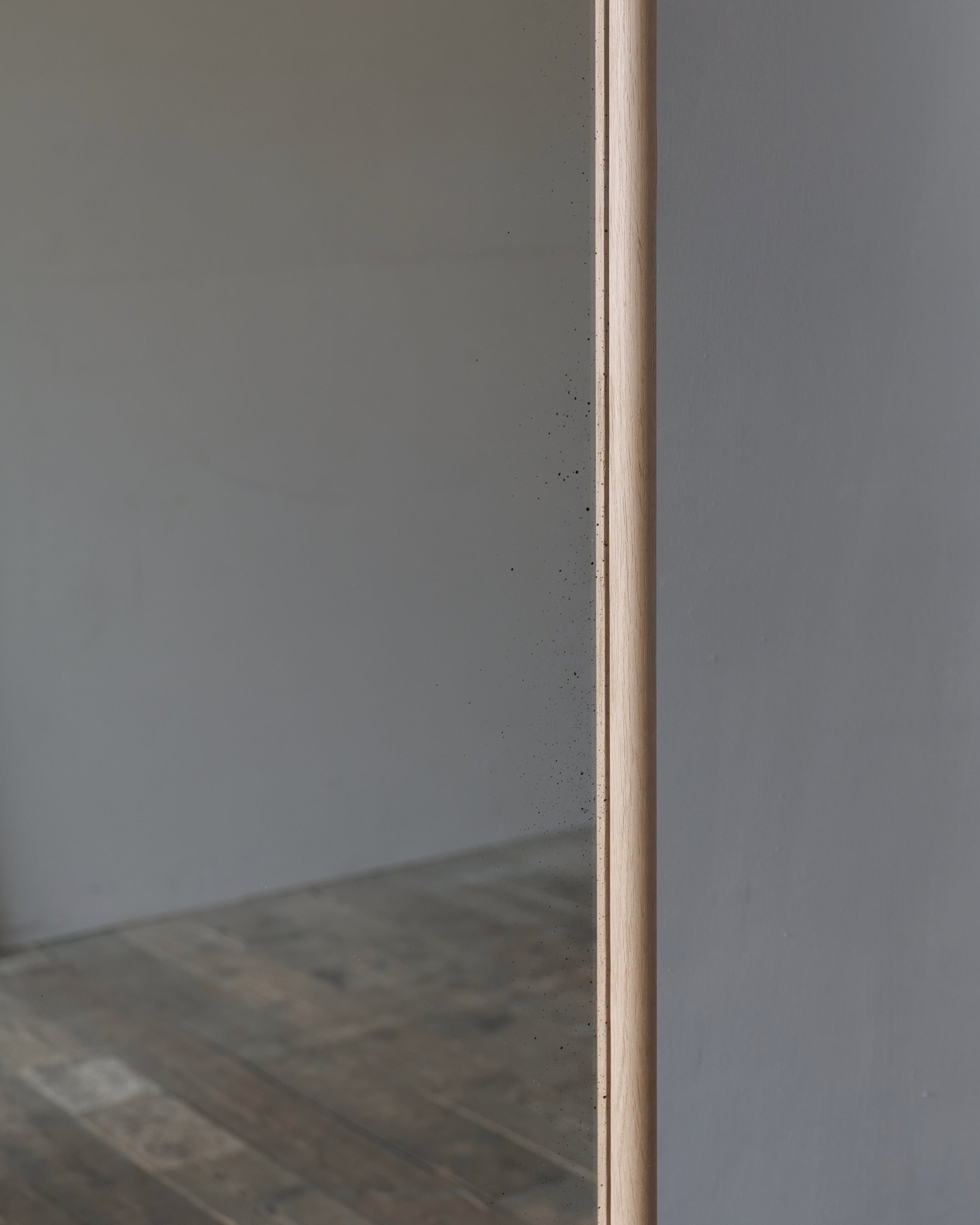 A close-up of a tall, narrow mirror with a light natural wood frame, reflecting a minimalist gray wall and rustic wooden floor.