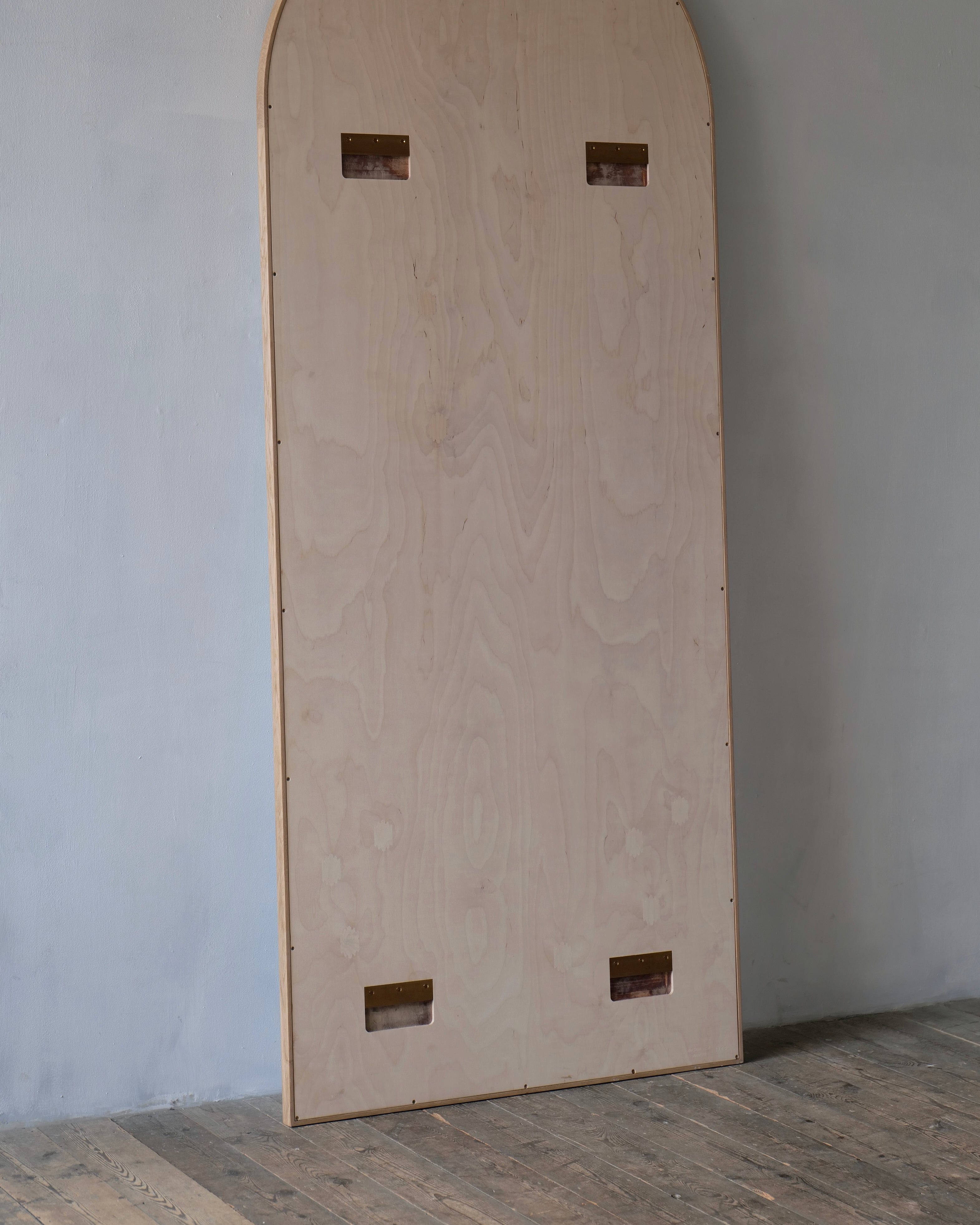 A tall, narrow light wood panel with rounded top corners and four rectangular cutouts, leaning against a plain wall on a wooden floor.