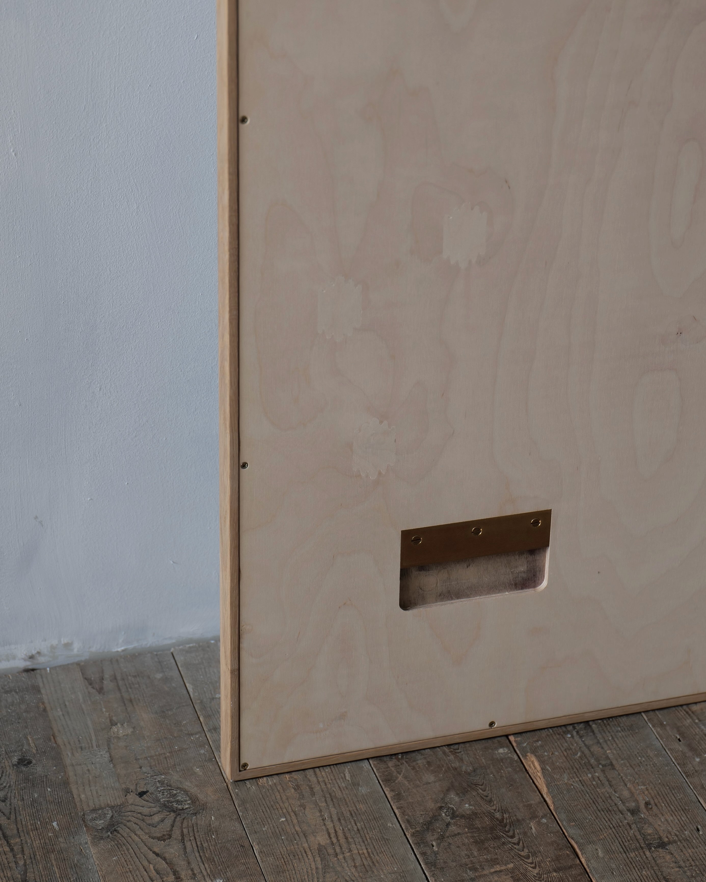 A large light-colored plywood panel standing on a rustic wooden floor against a white wall, with a rectangular metal cutout near the bottom.