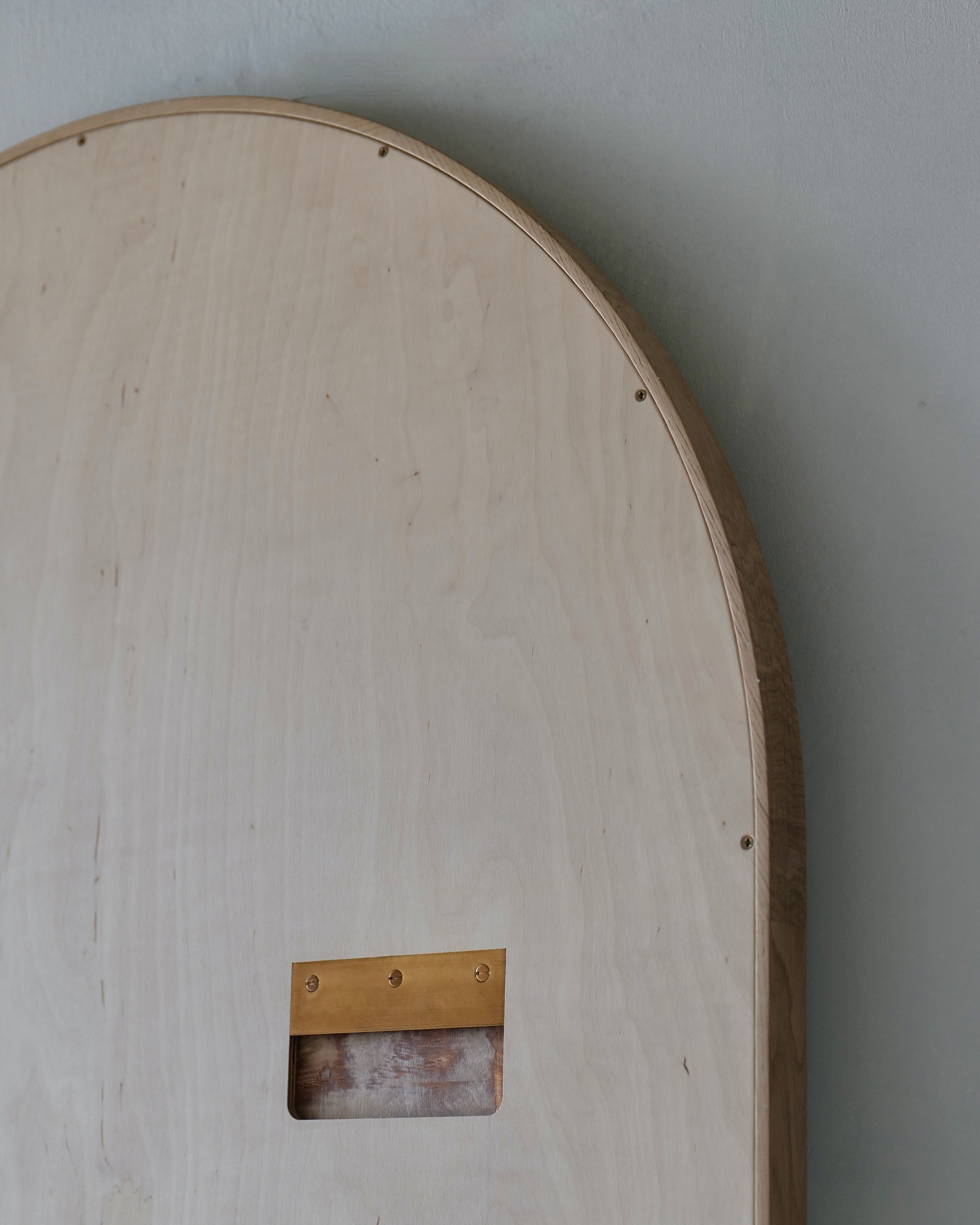 Close-up of a light natural wood arch-shaped panel or board with a rectangular cutout and a small brass-colored metal bracket, set against a plain gray wall.