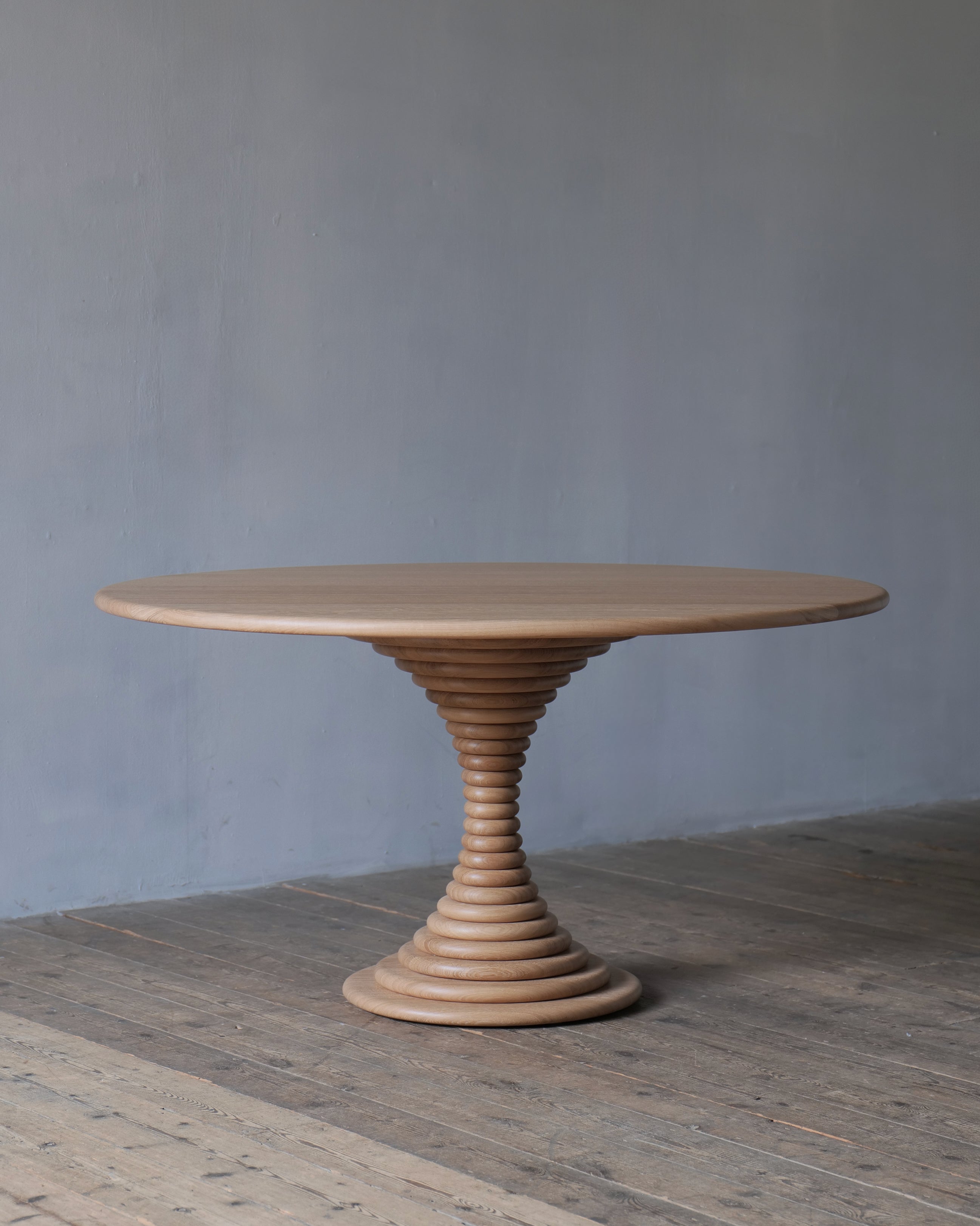 A circular natural oak socle dining table with an hourglass, pedestal base, set against a textured grey wall and rustic wooden floor.