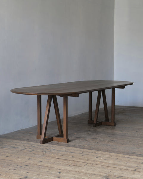An oval oak legacy dining table with a dark fumed finish and A-frame trestle legs, set in a minimalist room with a concrete wall and wooden floor.