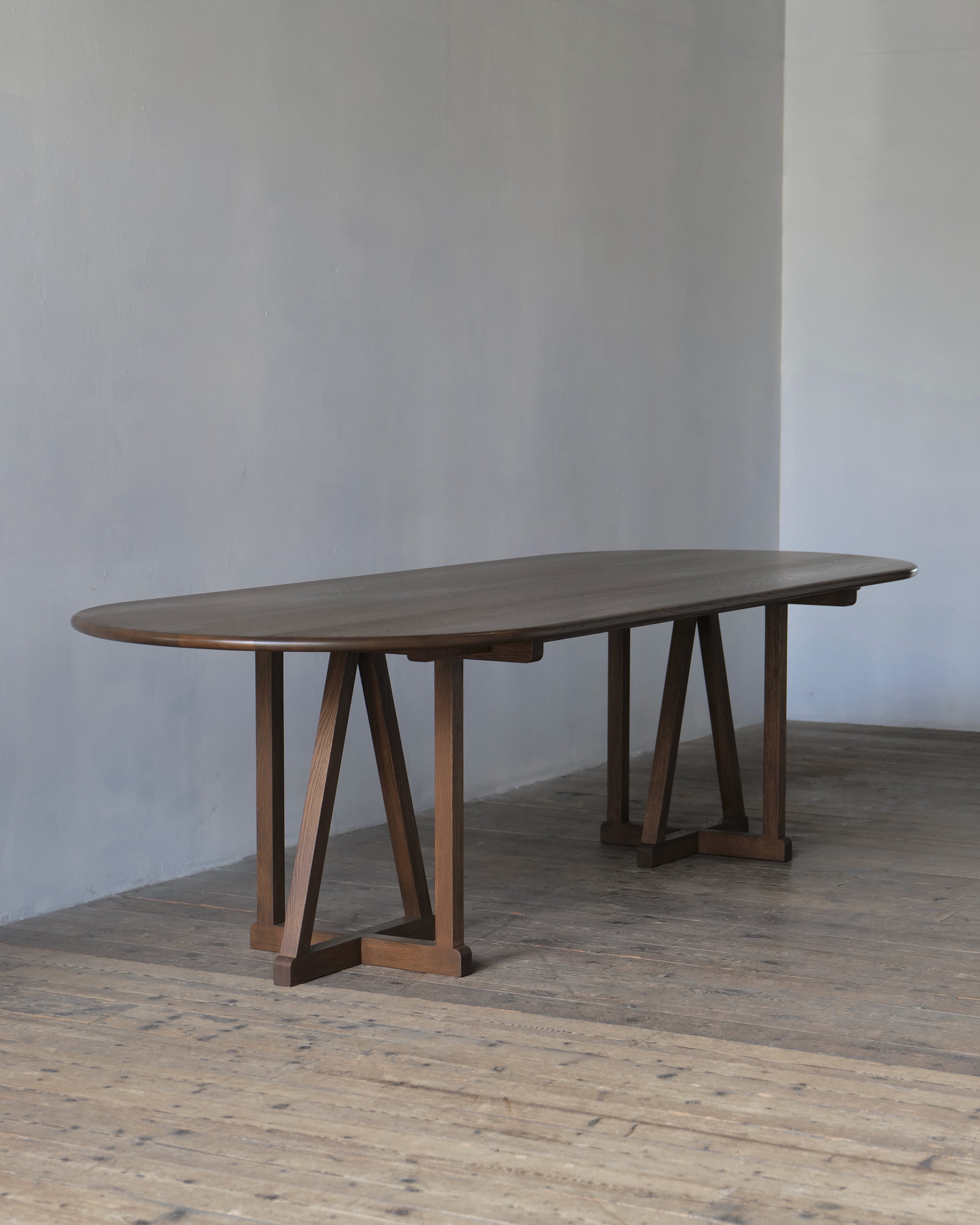 An oval oak legacy dining table with a dark fumed finish and A-frame trestle legs, set in a minimalist room with a concrete wall and wooden floor.