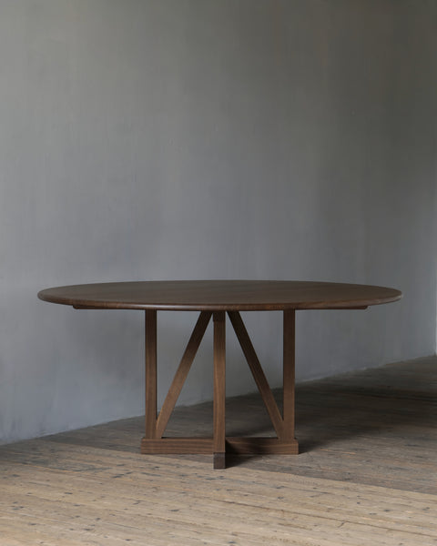 A circular legacy dining table with a dark fumed finish and a central trestle-style base, set against a textured grey wall and rustic wooden floor.