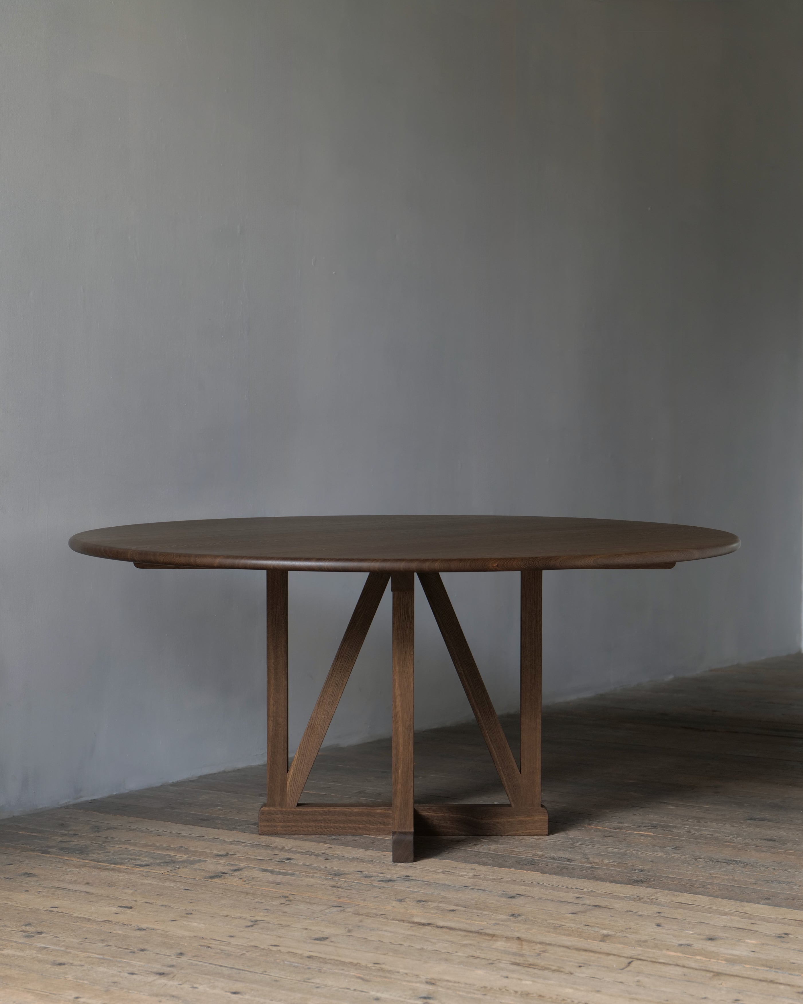 A circular legacy dining table with a dark fumed finish and a central trestle-style base, set against a textured grey wall and rustic wooden floor.