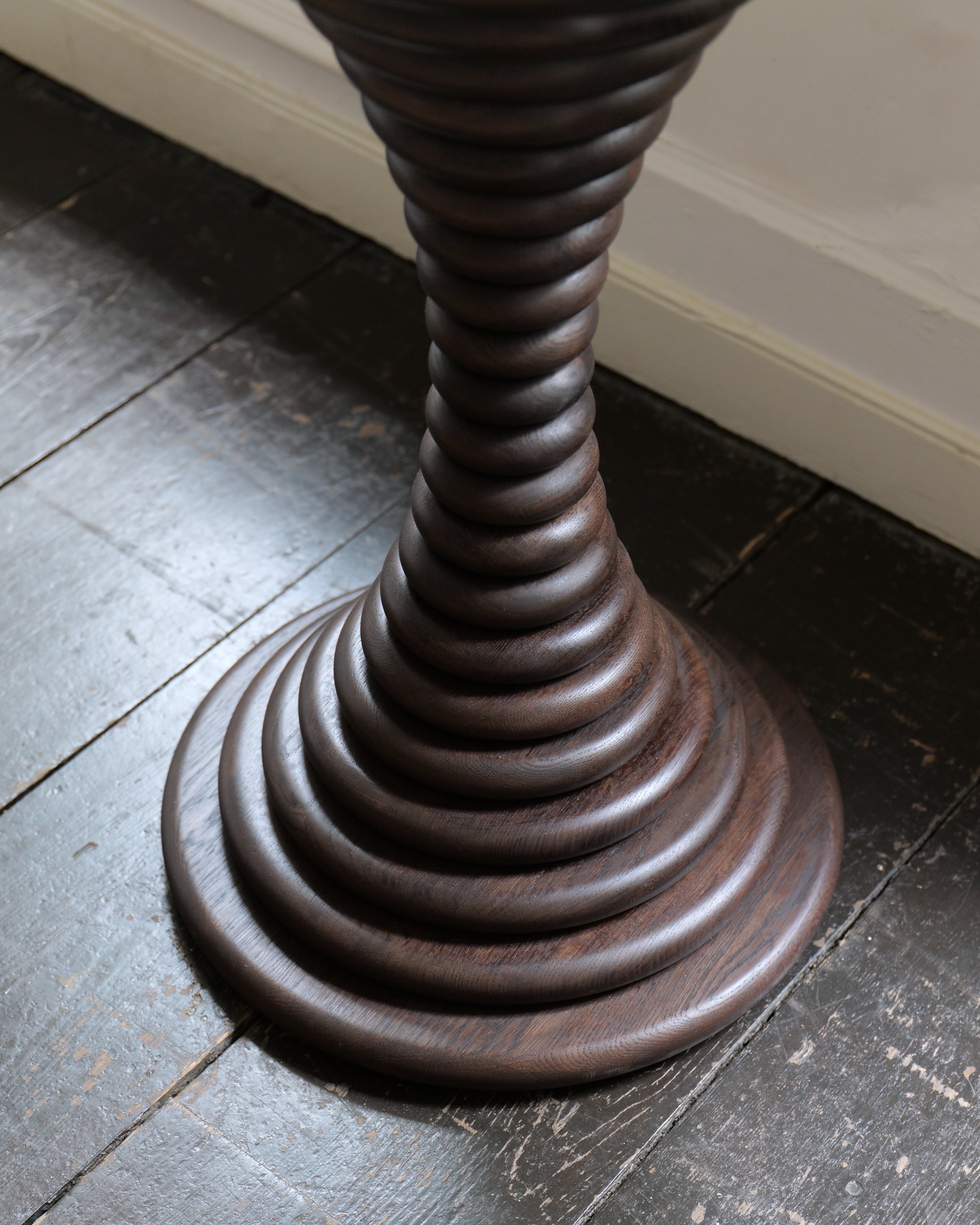 A close-up of a dark brown wooden pedestal table base, set on a dark wood floor against a white wall.