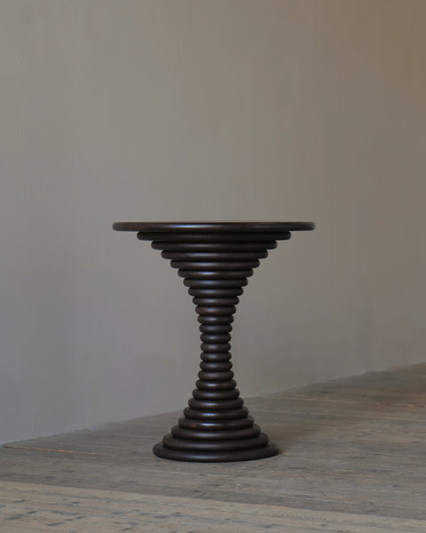 A dark brown wooden pedestal table with a circular top, set against a neutral beige wall and textured grey floor.